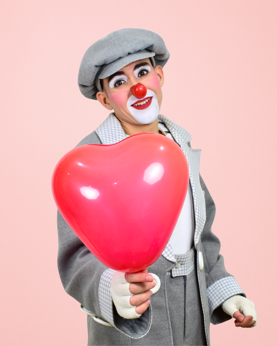 Payaso con maquillaje, sombrero gris, y ropa a cuadros sosteniendo un globo en forma de corazón rosa, frente a un fondo rosa.