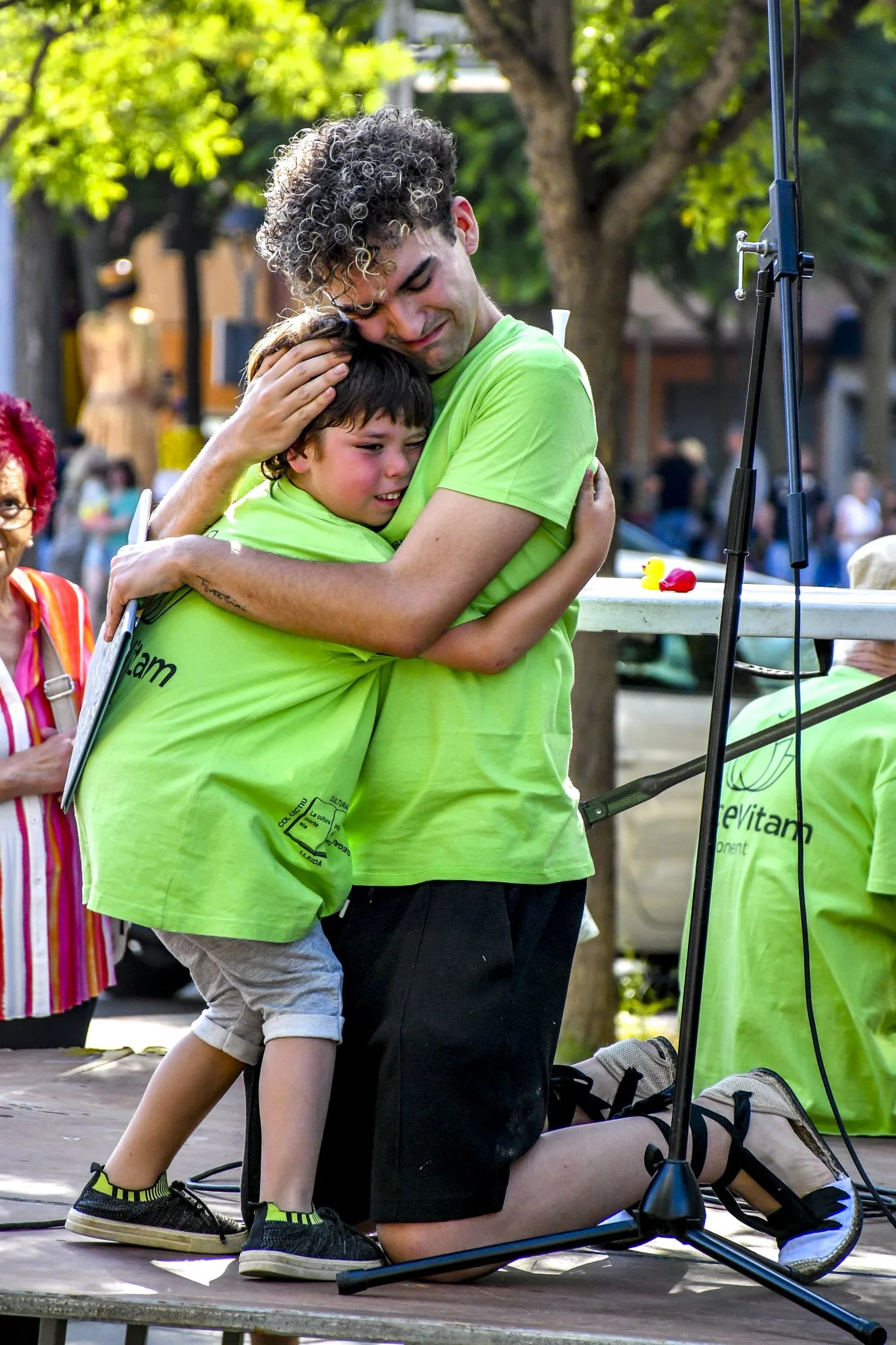 Un hombre y un niño pequeños se abrazan emotivamente en una plaza, mientras otros adultos observan. Ambos llevan camisetas verdes y parecen estar en un evento público.