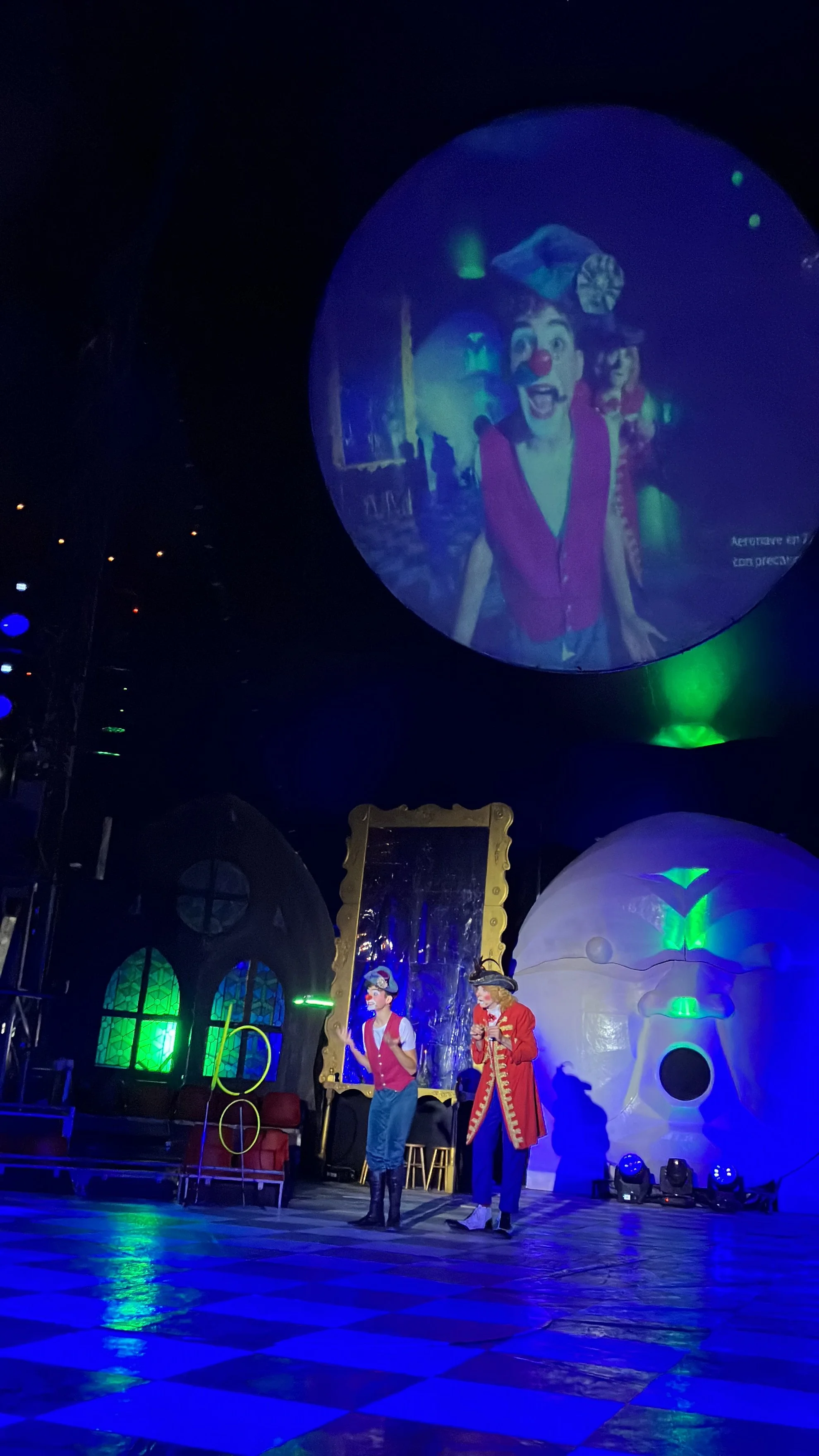 Espectáculo en teatro con dos actores en disfraces, escenario decorado con elementos coloridos y un proyector gigante mostrando una imagen de payasos en el cielo oscuro.