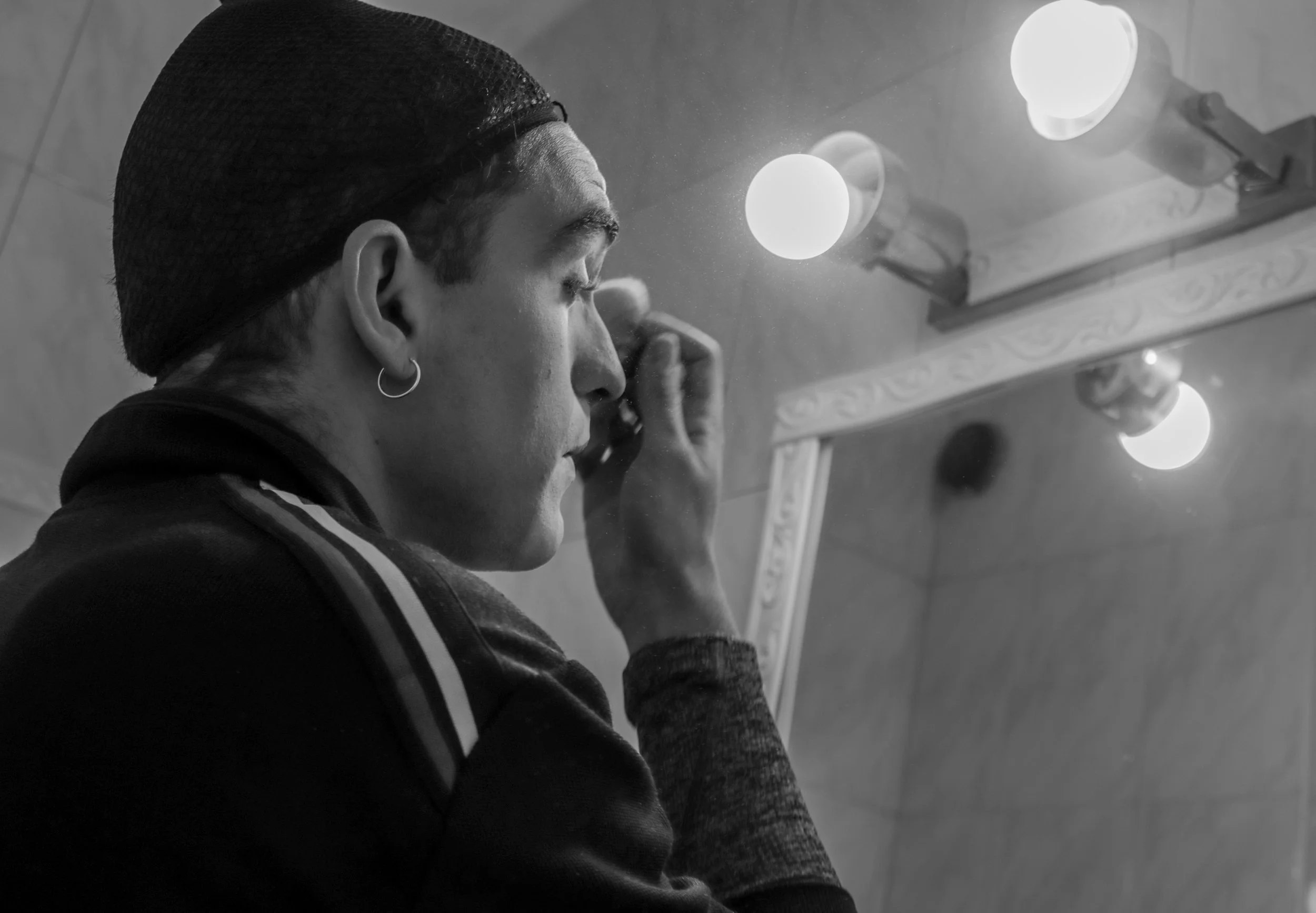 Una persona en un camarín, arreglándose el maquillaje frente a un espejo con luces, en blanco y negro.