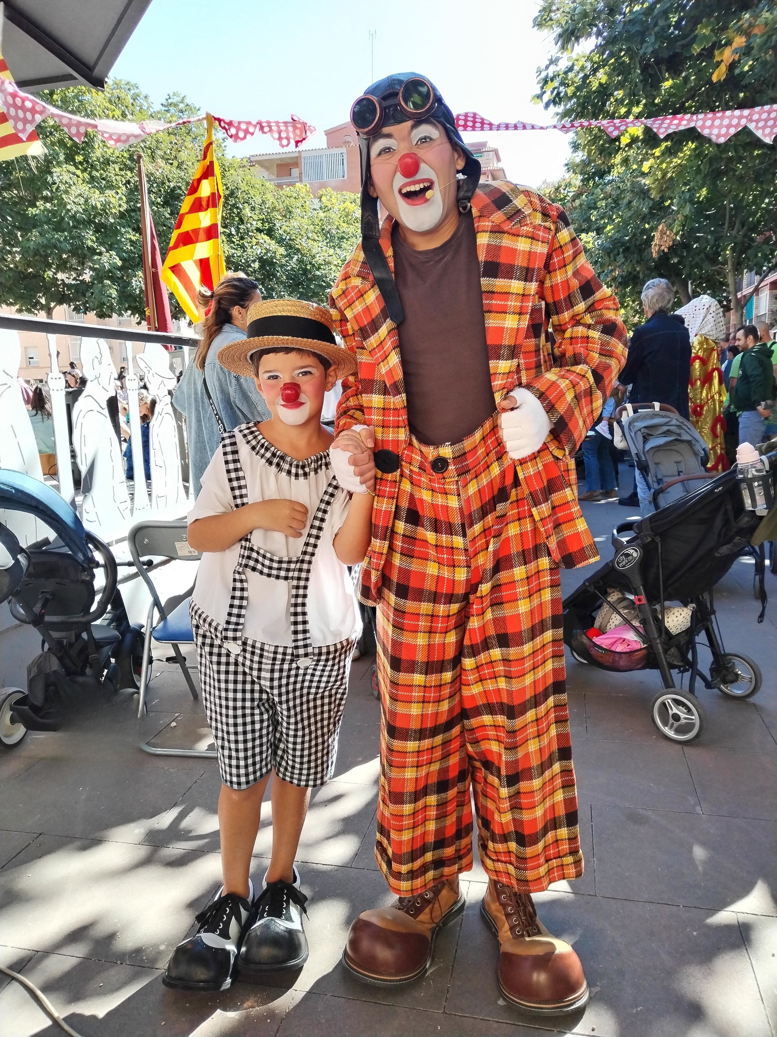 Niño y payaso disfrazados de payasos con narices rojas en una celebración en exteriores, con banderas y demás personas en el fondo.