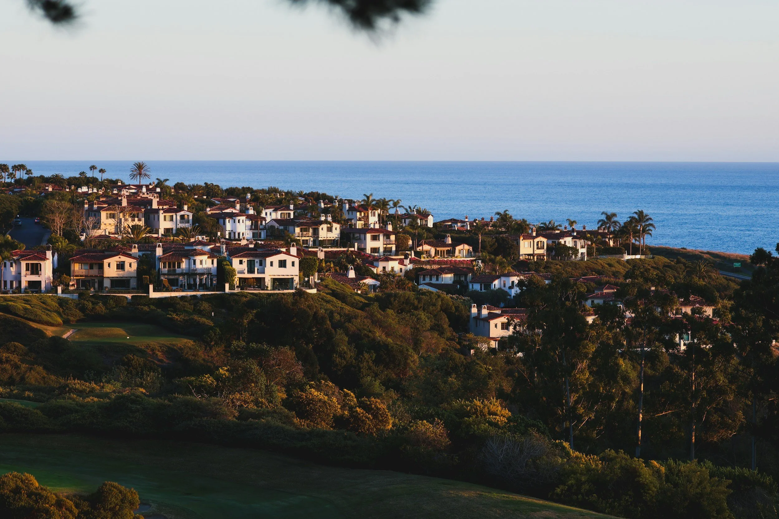 A scenic view of a coastal residential neighborhood with white houses featuring red-tiled roofs, lush green trees, and a golf course in the foreground, overlooking the ocean in the background.