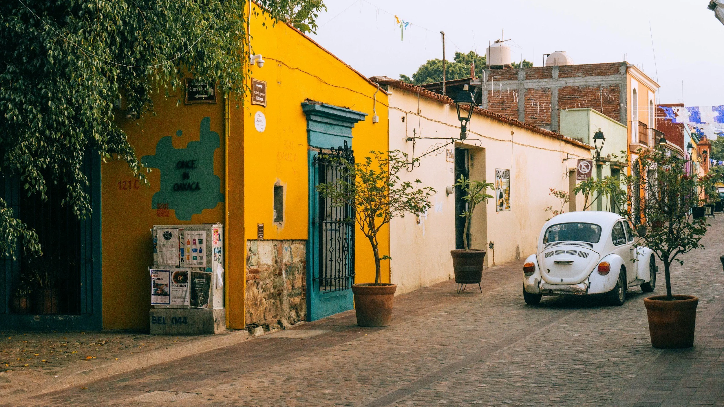 Colorful buildings with yellow, blue, green, and red walls line a cobblestone street, with potted plants and a white vintage car parked along the sidewalk. Decorative street lamps and hanging banners are visible against a clear sky.