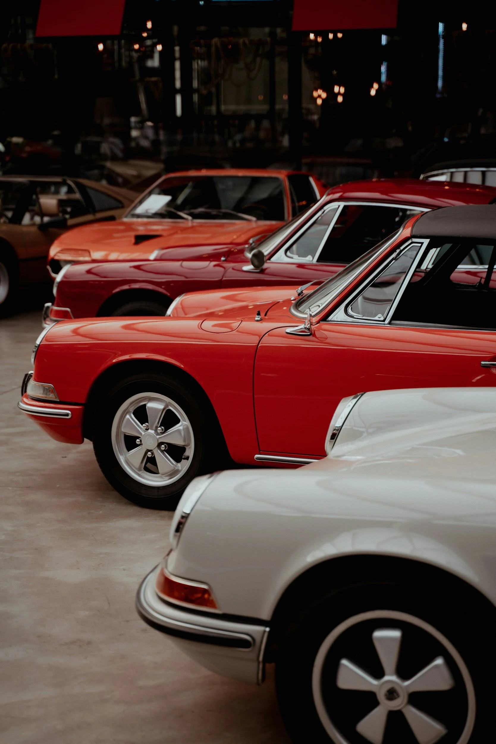 Multiple vintage sports cars in red, orange, and white parked indoors at a classic car show or museum.
