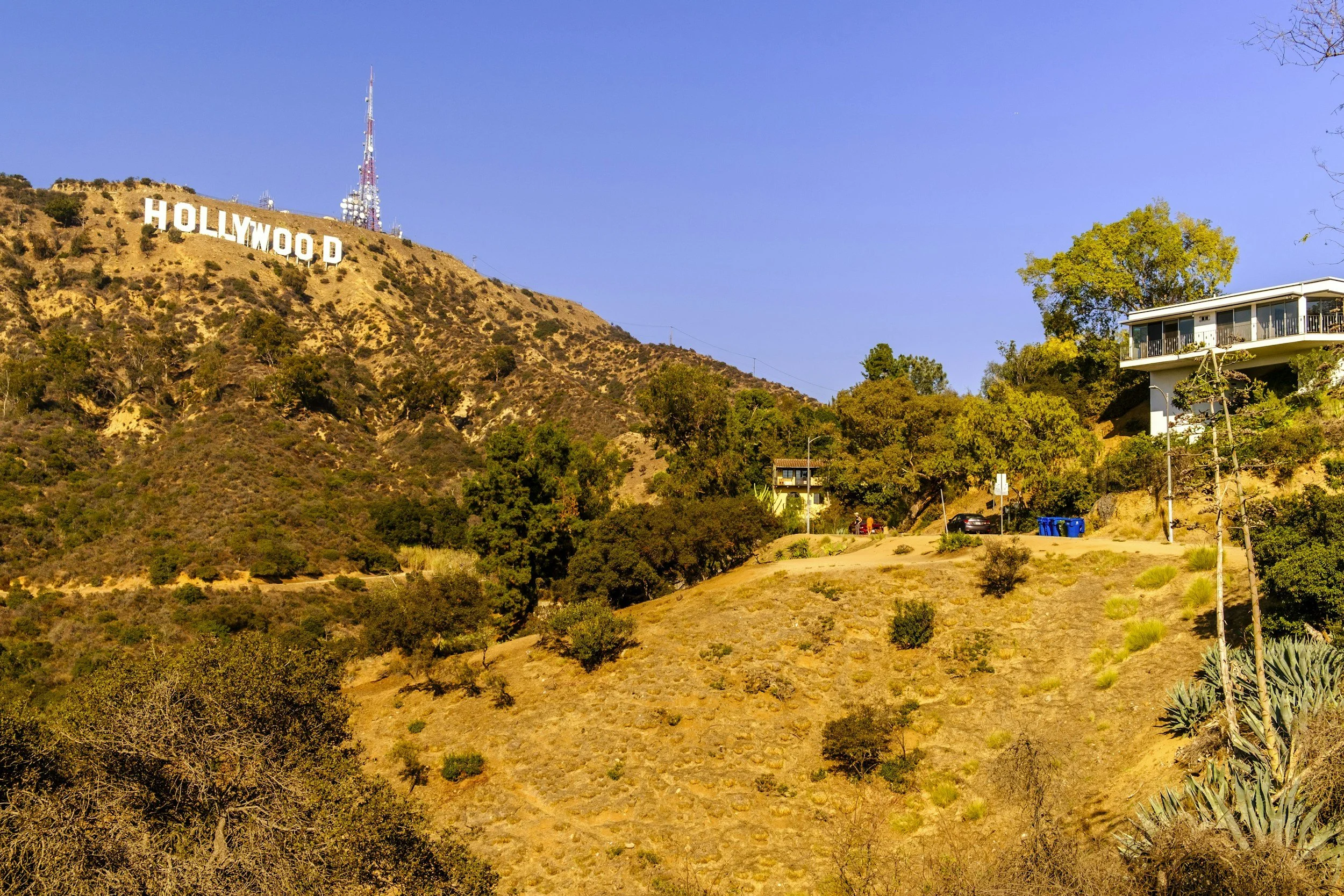 The Hollywood sign on a hillside with dry vegetation, trees, a house on the right, and a blue sky.