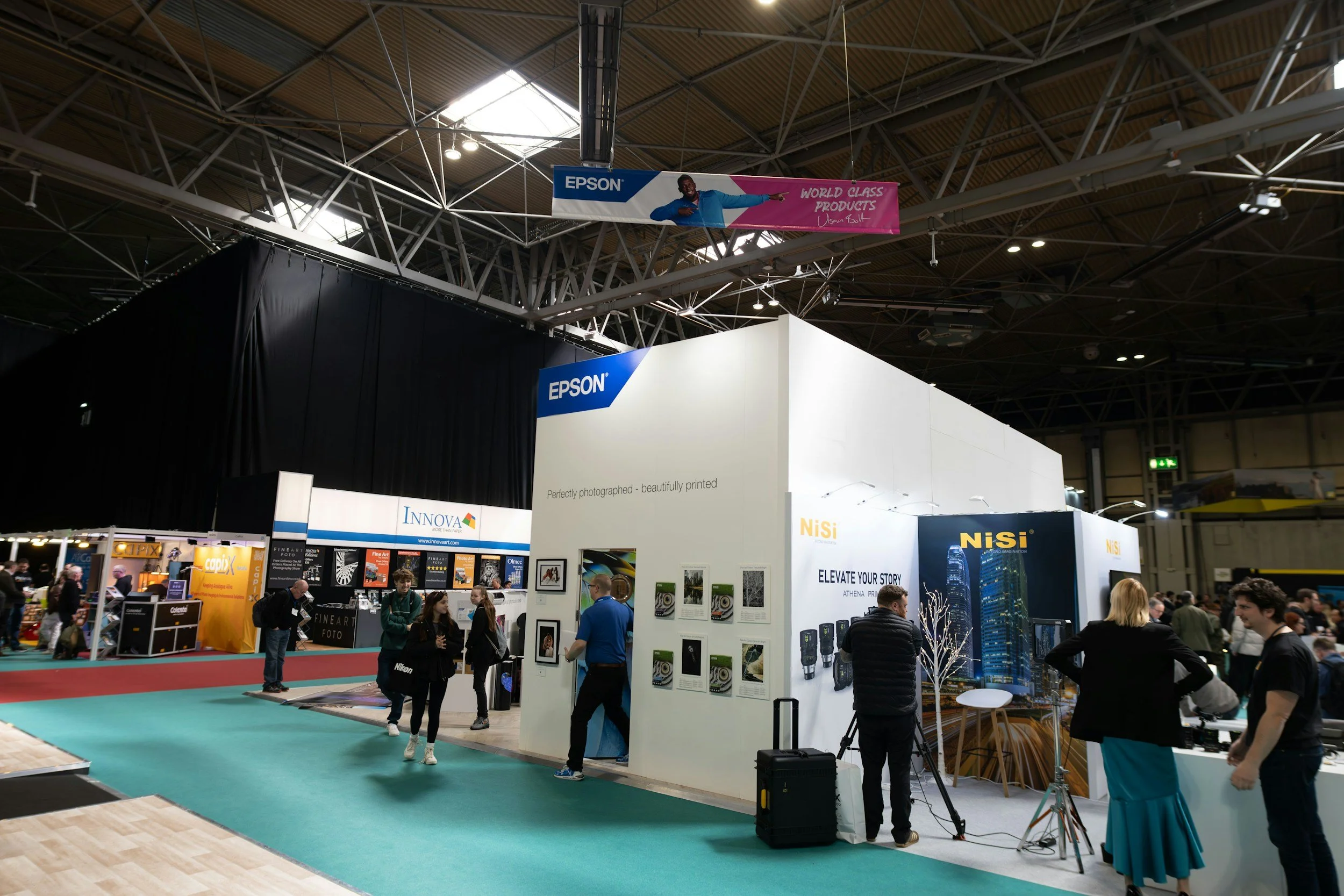 A busy trade show or exhibition with various booths and displays. People are walking around and engaging with exhibitors. A large banner with the Epson logo and a person pointing is hanging from the ceiling. The floor has a mix of carpeted and wooden areas, and the ceiling is high with visible metal trusses.
