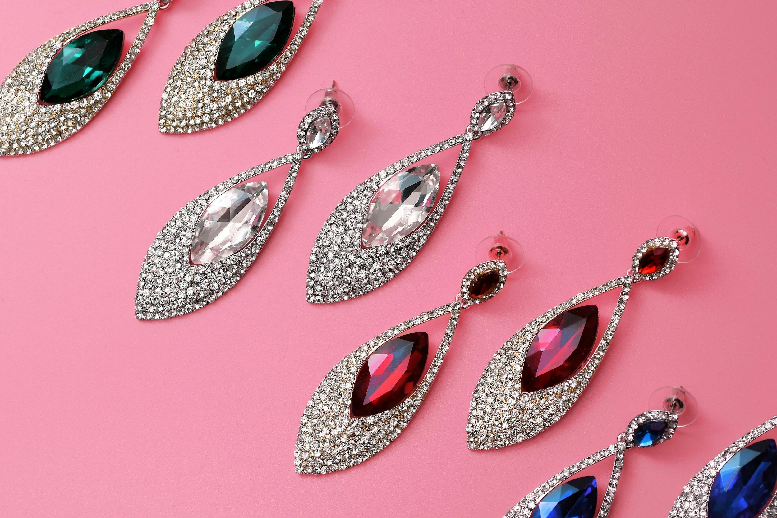 Pair of large, ornate earrings with sparkling clear and green gemstones on a pink background.