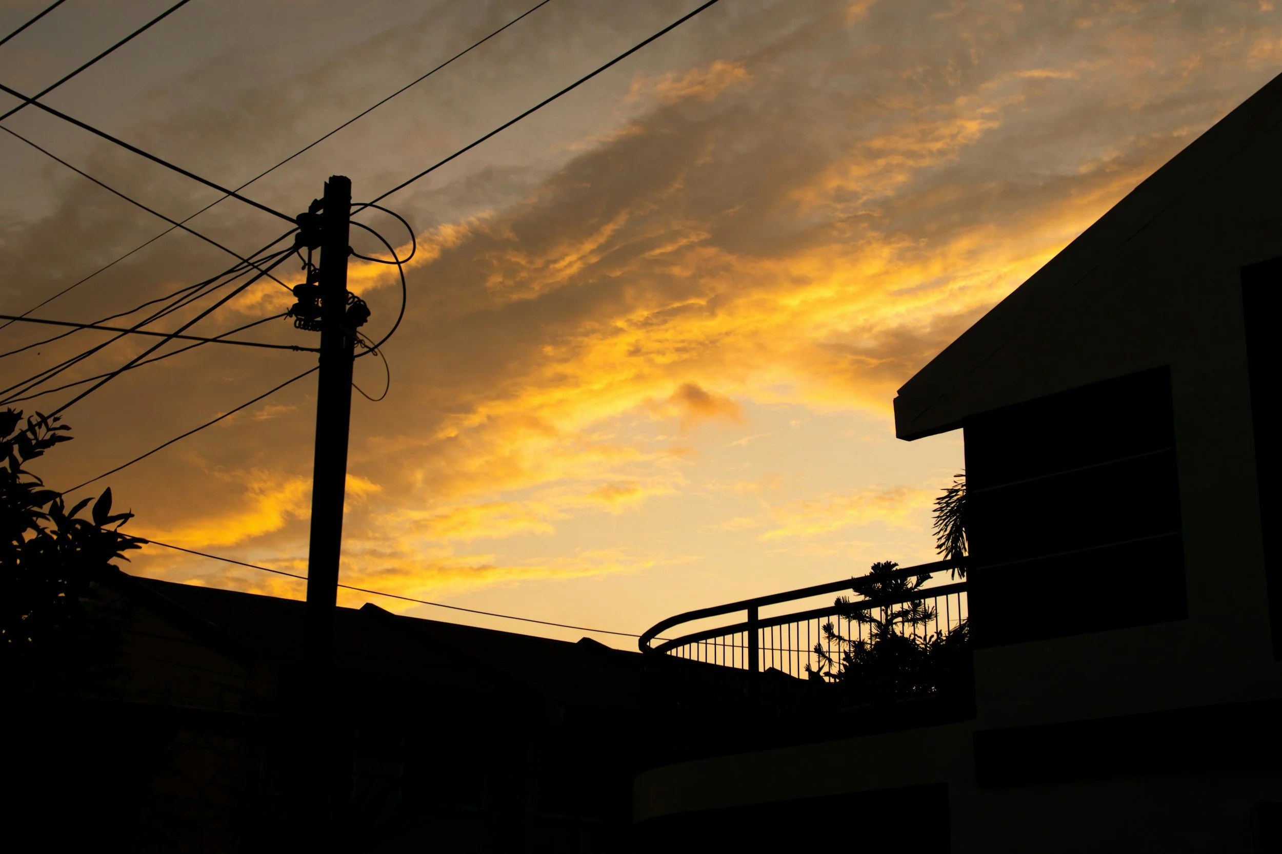 Silhouette of a utility pole with wires against a sunset sky with orange and yellow clouds, partly obscured by the building and trees in the foreground.