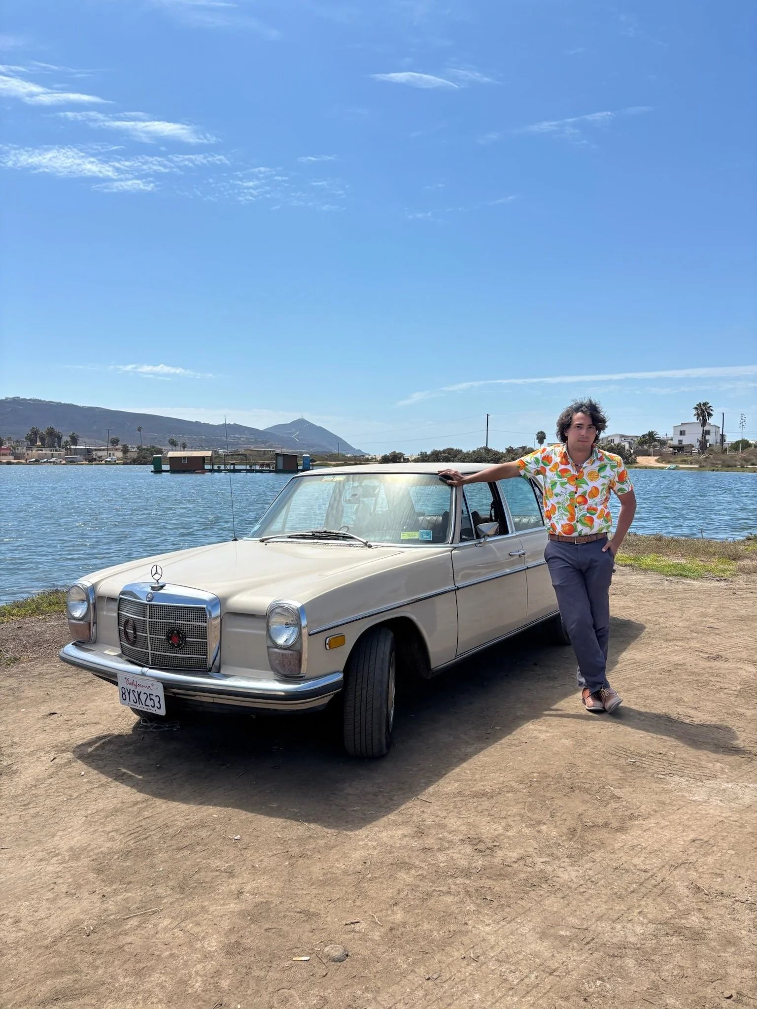 A man in a colorful Hawaiian shirt and gray pants standing next to a vintage beige Mercedes-Benz car by a water body with hills and residential buildings in the background.