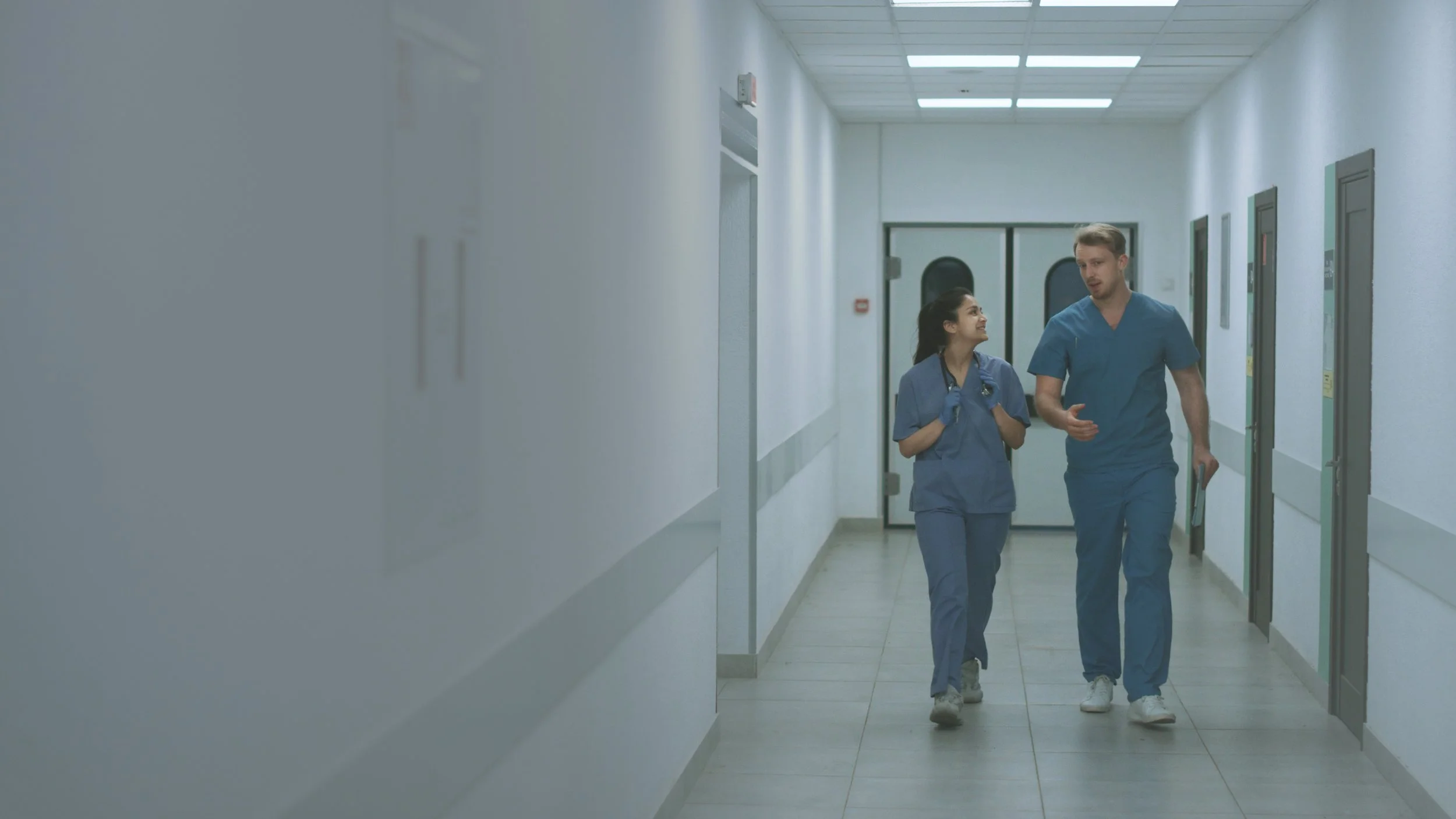 Two healthcare providers in scrubs walking and conversing in a clean, secure institutional hallway.