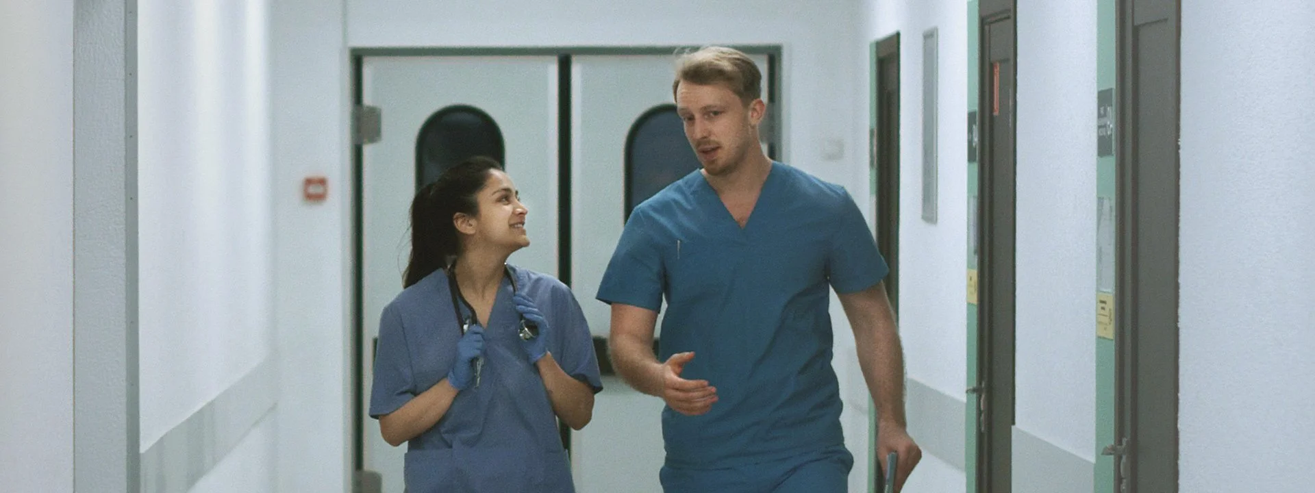 Two healthcare professionals in blue scrubs walking and conversing in a brightly lit, modern clinical hallway.