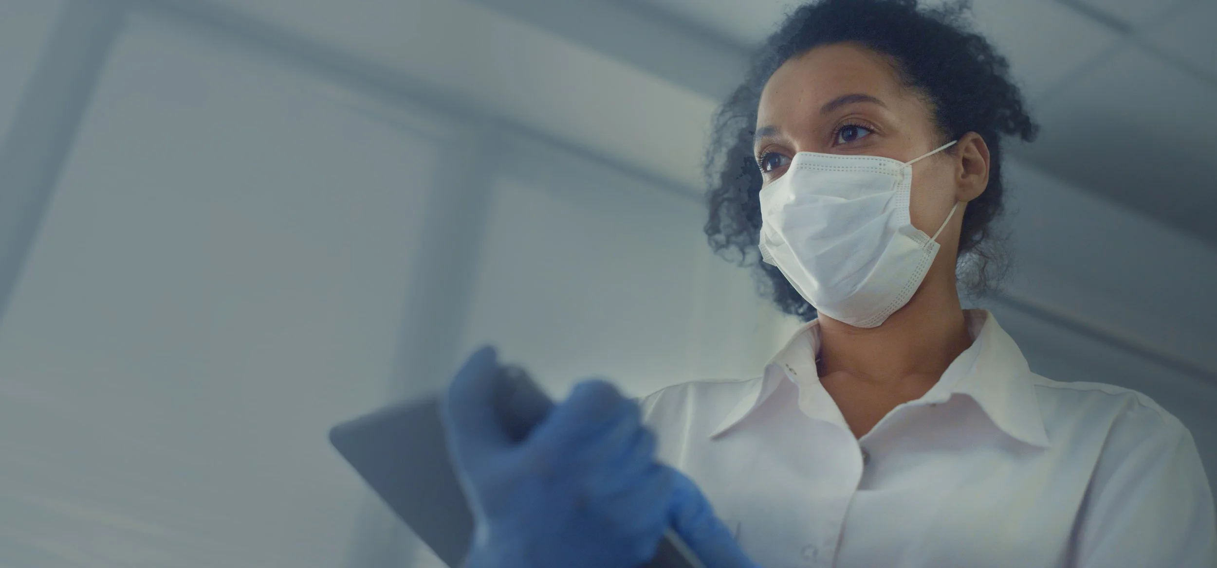 A female healthcare provider wearing a surgical mask and white coat, holding a tablet and looking thoughtfully forward.