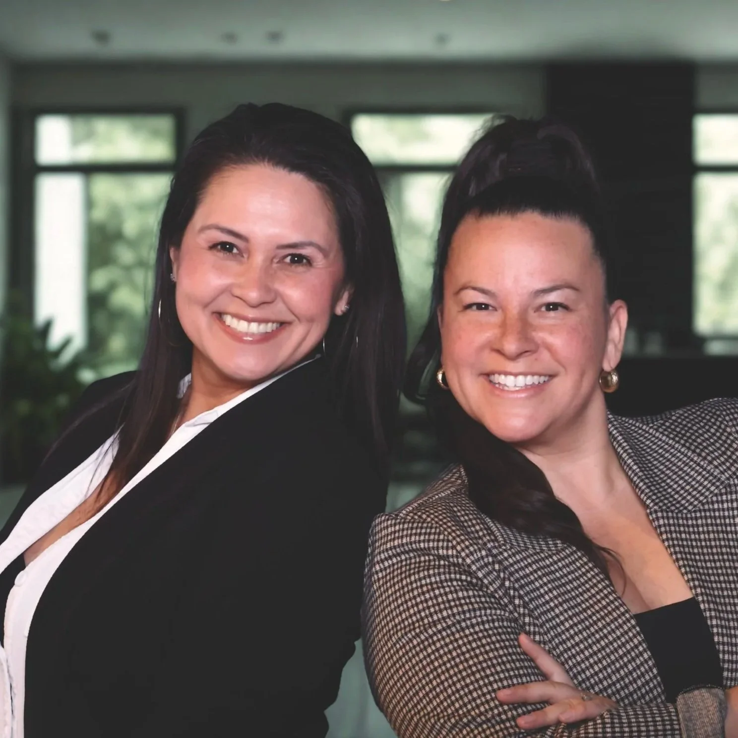 Two women smiling and posing for a photo indoors, with large windows and greenery in the background.