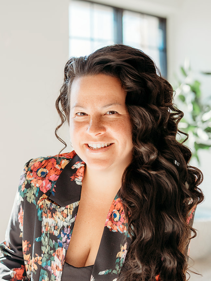Smiling woman with long, curly dark hair wearing a colorful floral blazer indoors.