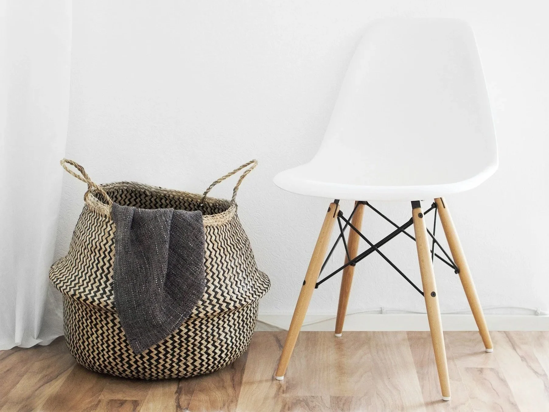 A woven basket filled with dark-colored cloths, sitting on a hardwood floor next to a white modern chair with wooden legs.