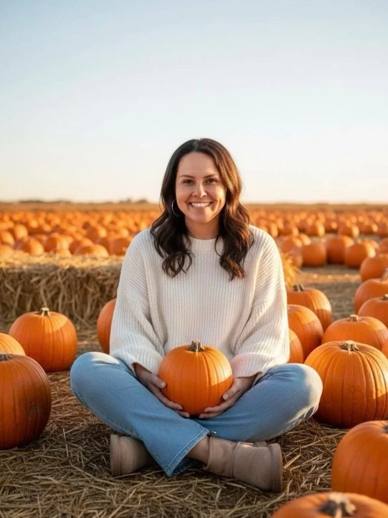 A woman sitting on the ground in a pumpkin patch holding a small pumpkin, surrounded by many other pumpkins under a clear sky.