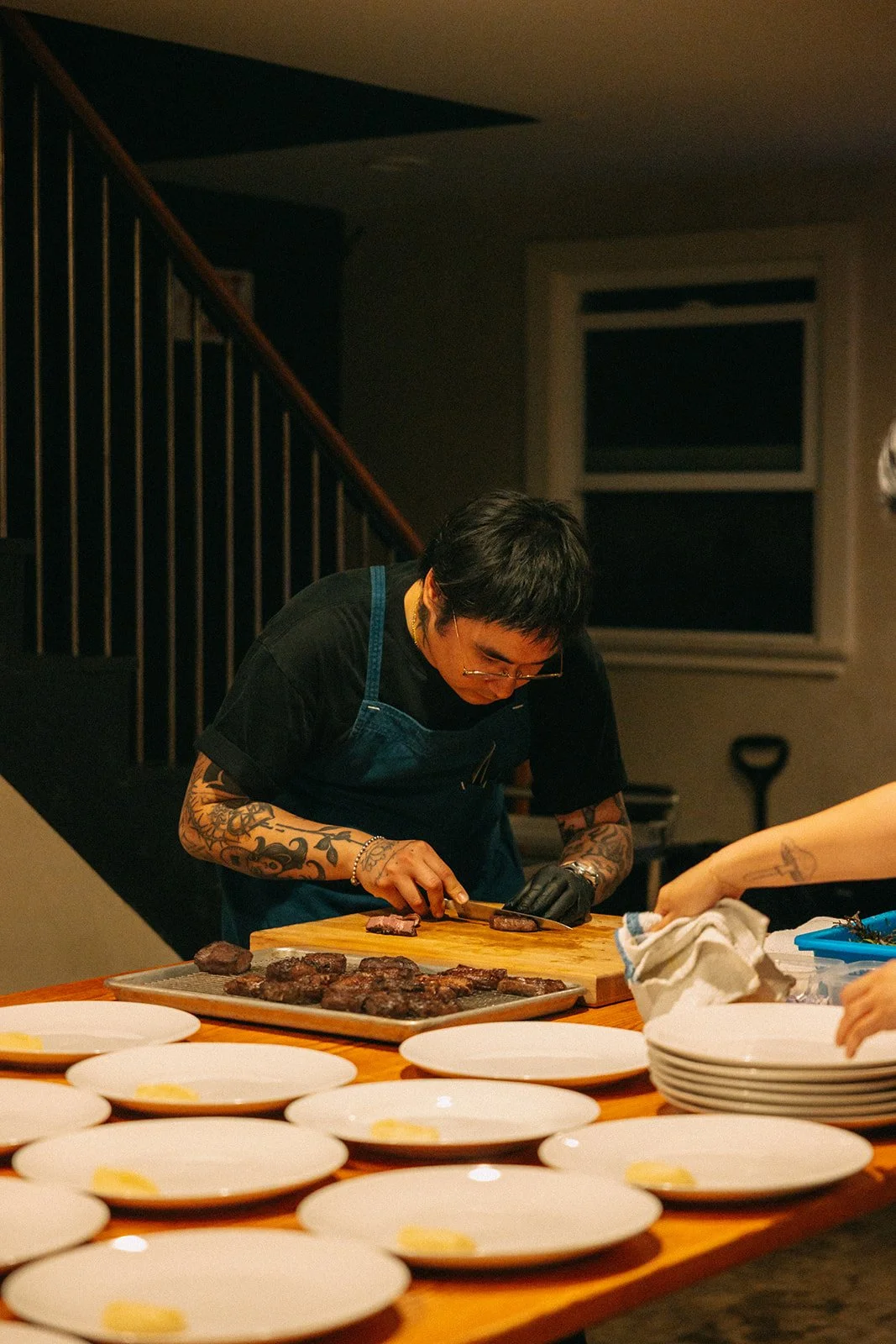 Person with tattoos preparing grilled steaks on a wooden cutting board in a kitchen.