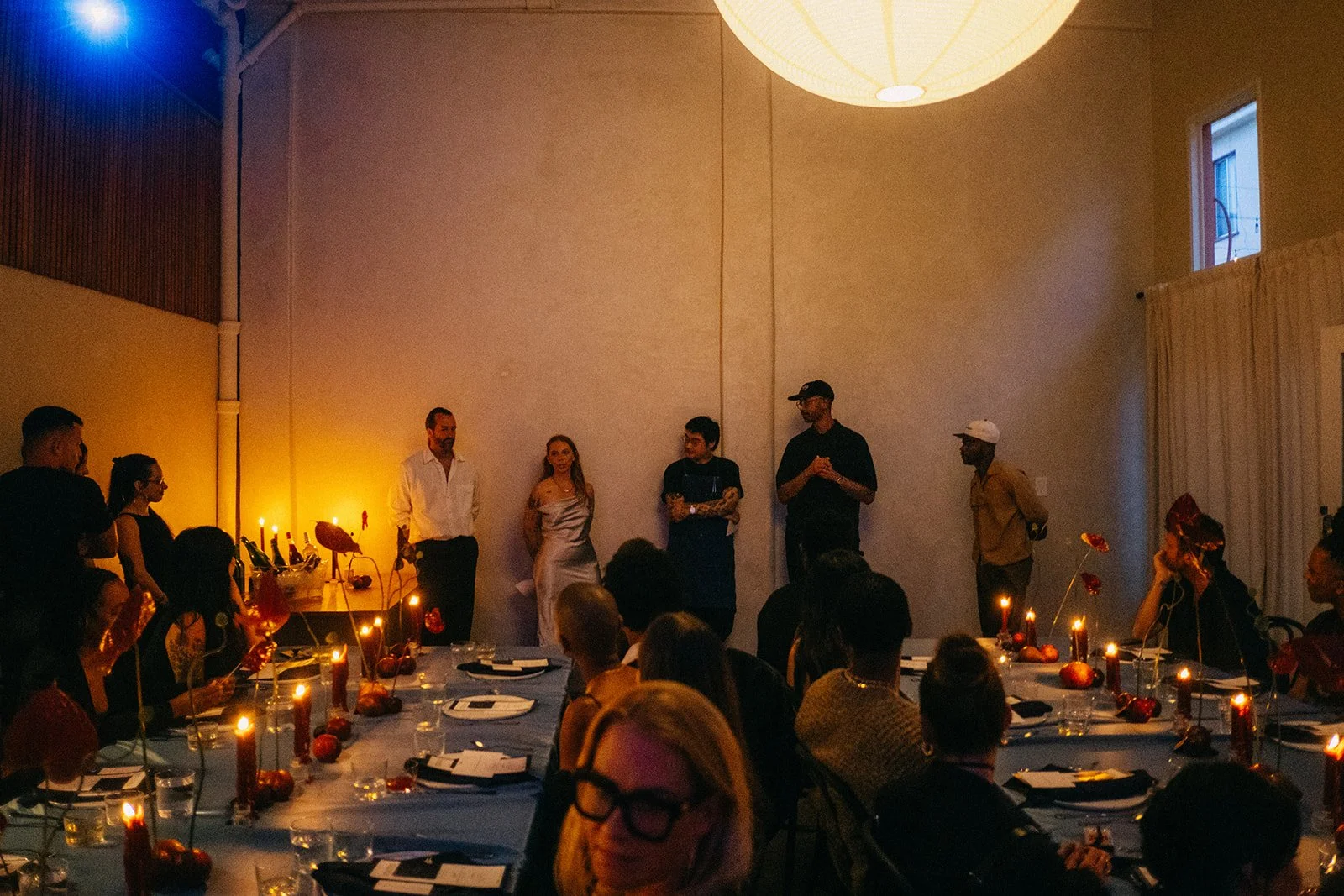 People gathered in a dimly lit room with a high ceiling, watching a speaker on stage. The room has round tables with candles, plates, and glasses, and the audience is seated and attentively listening.