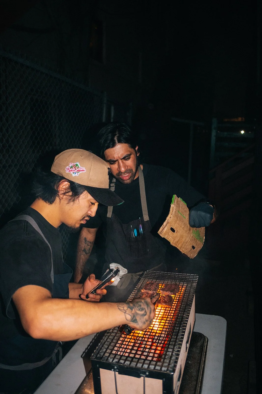 Two men grilling meat over an open flame at night, one is using a smartphone and the other is holding a piece of cardboard.