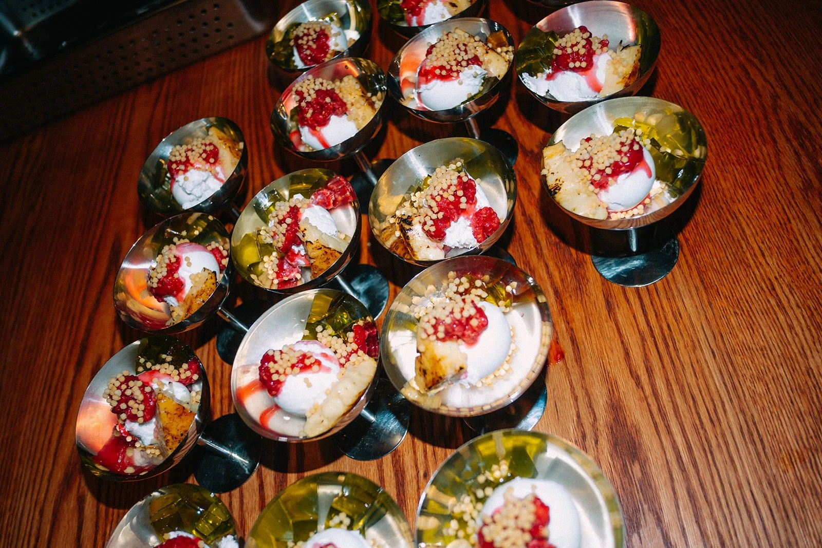 Multiple small dessert cups on a wooden surface, each filled with whipped cream, red syrup, yellow gelatin, white pearls, and crumbled cookie pieces.