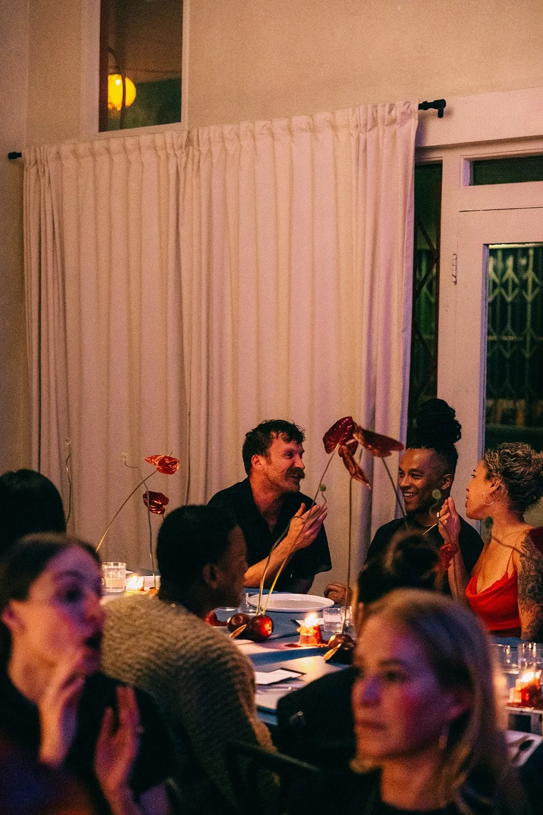 People sitting around a dinner table at a social gathering, smiling and engaging in conversation, with a curtain in the background and small decorations on the table.