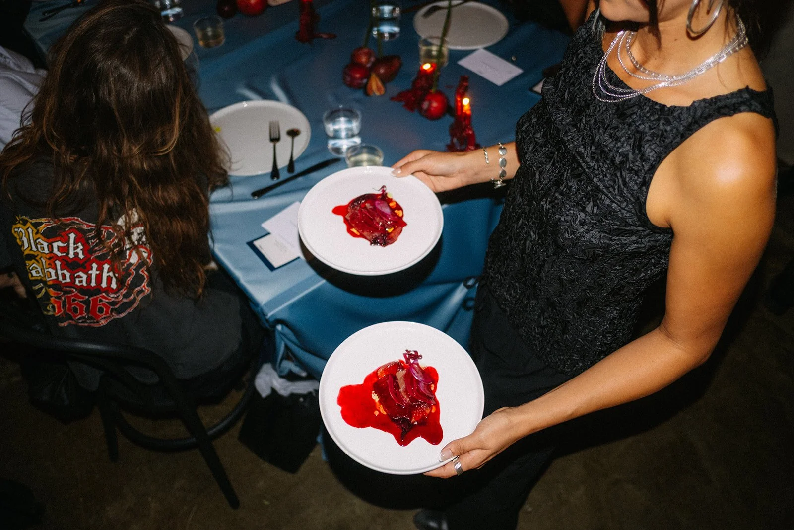 Person holding two white plates with sliced meat drizzled with red sauce at a blue-covered dinner table decorated with candles and apples.