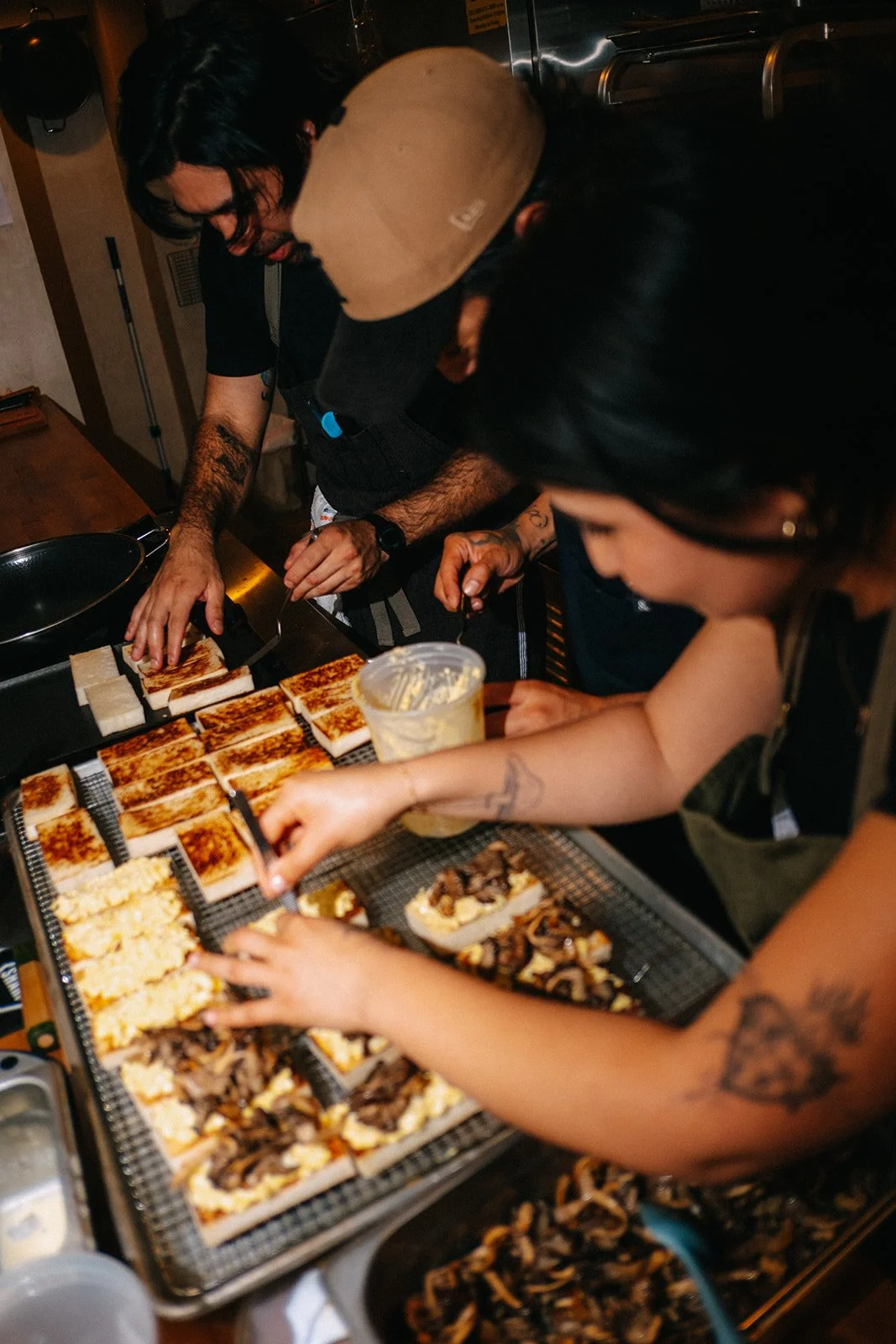 Two people preparing pizza slices in a kitchen. One person is placing toppings on pizza, while the other is cutting or arranging slices on a tray. There are toppings and ingredients on the counter, including cheese and mushrooms.