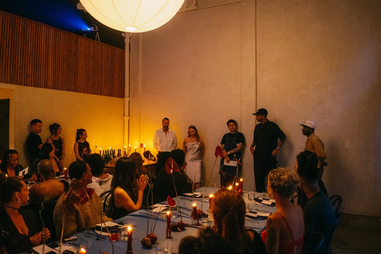 Guests seated at tables in a dimly lit restaurant or banquet hall, watching people stand near a lit bar against a yellow-orange wall, with some candles and decorations on the tables.