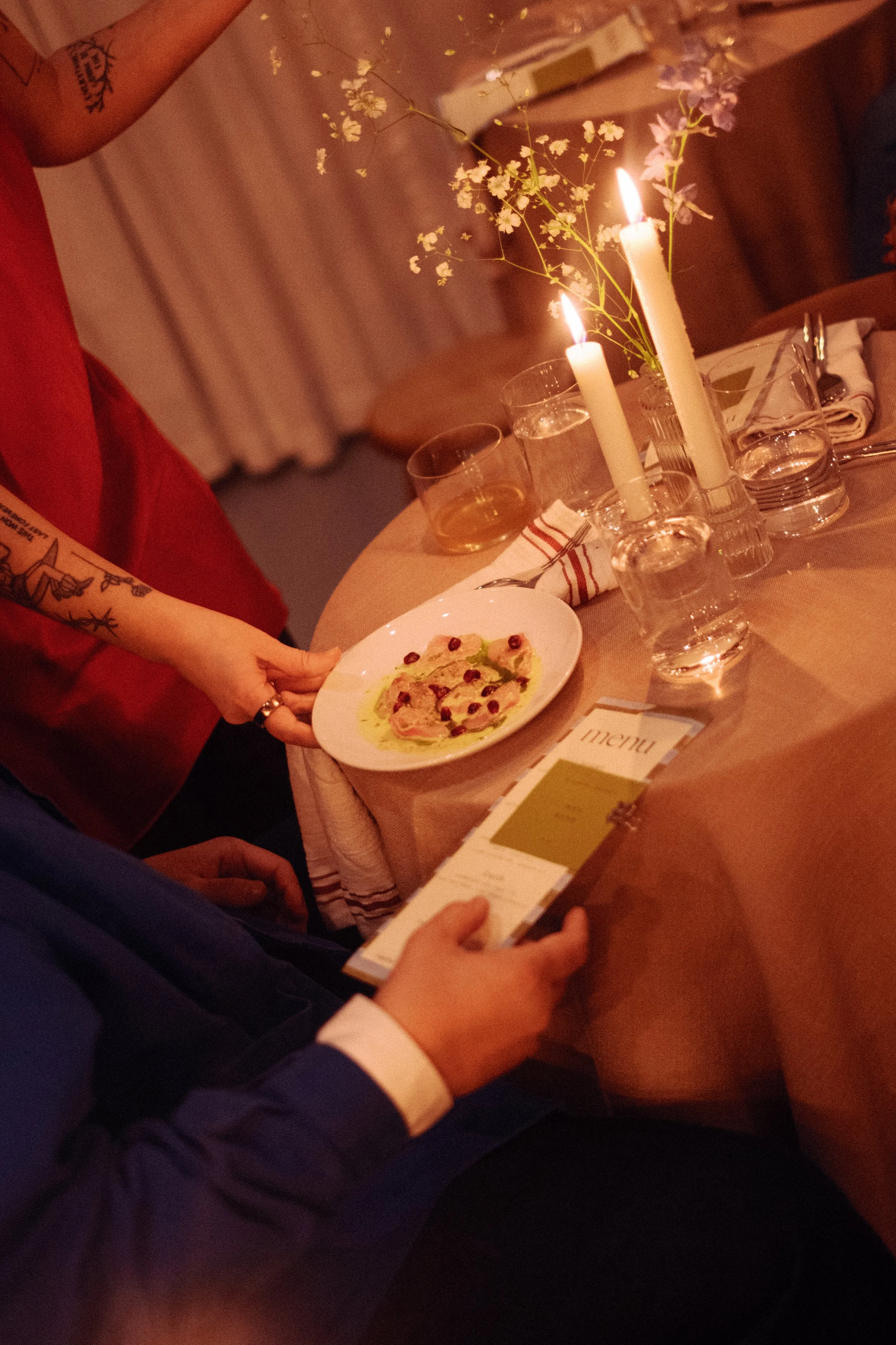 A dinner table with lit candles, a floral centerpiece, a menu, water glasses, a plate of food, and drinks. A person in a red shirt is holding a plate, and another person in a suit is holding a menu.