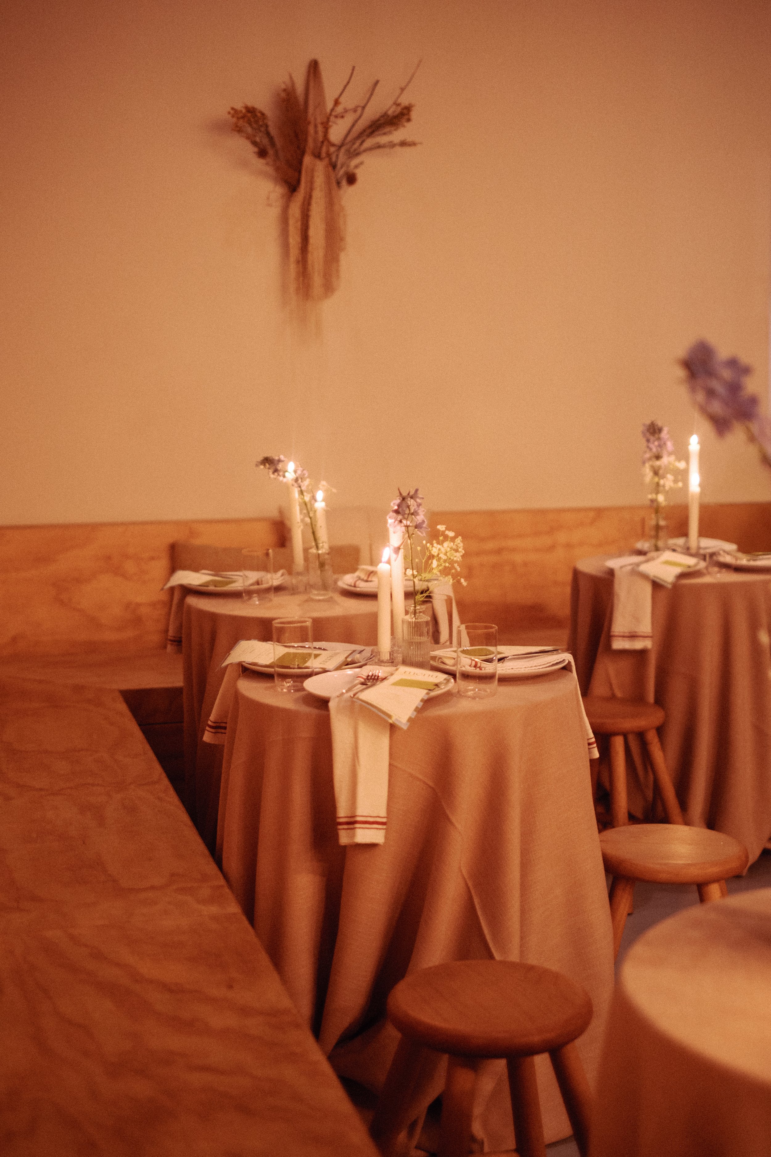 Dining room with three round tables set with candles, flowers, and place settings, surrounded by wooden benches and chairs, in a warm, cozy ambiance.