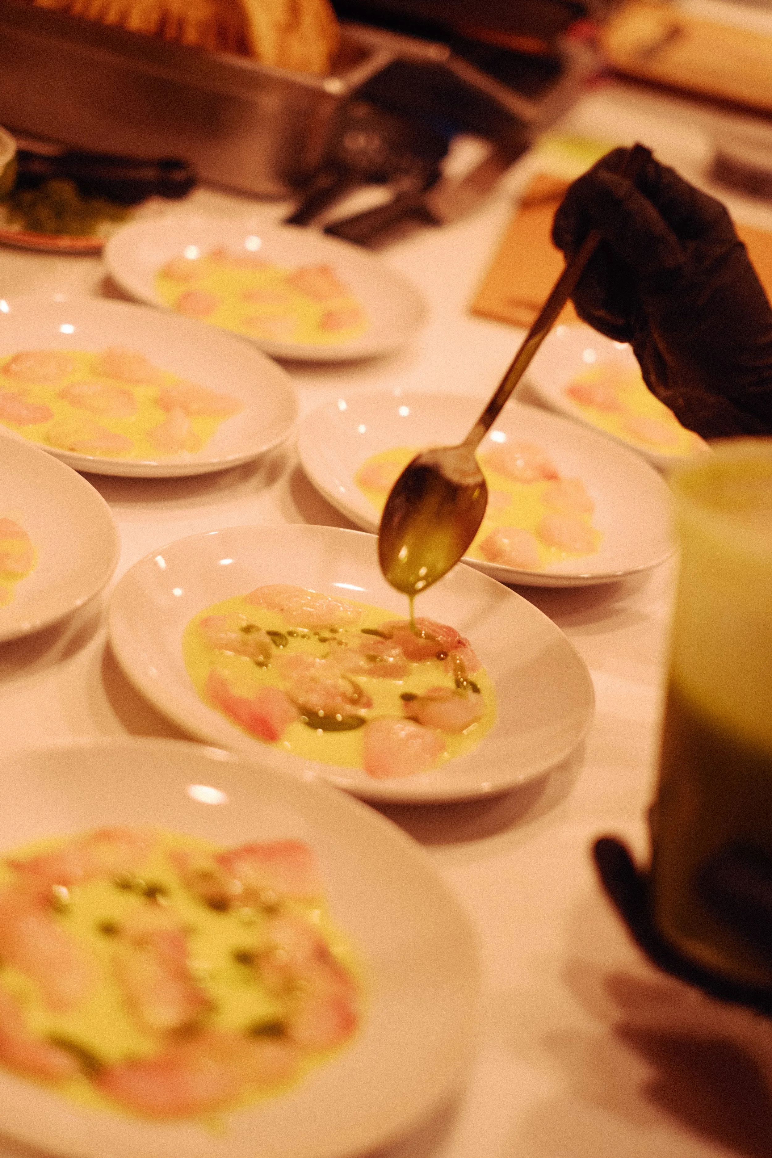 A chef drizzles olive oil over plates of raw shrimp in a professional kitchen setting.