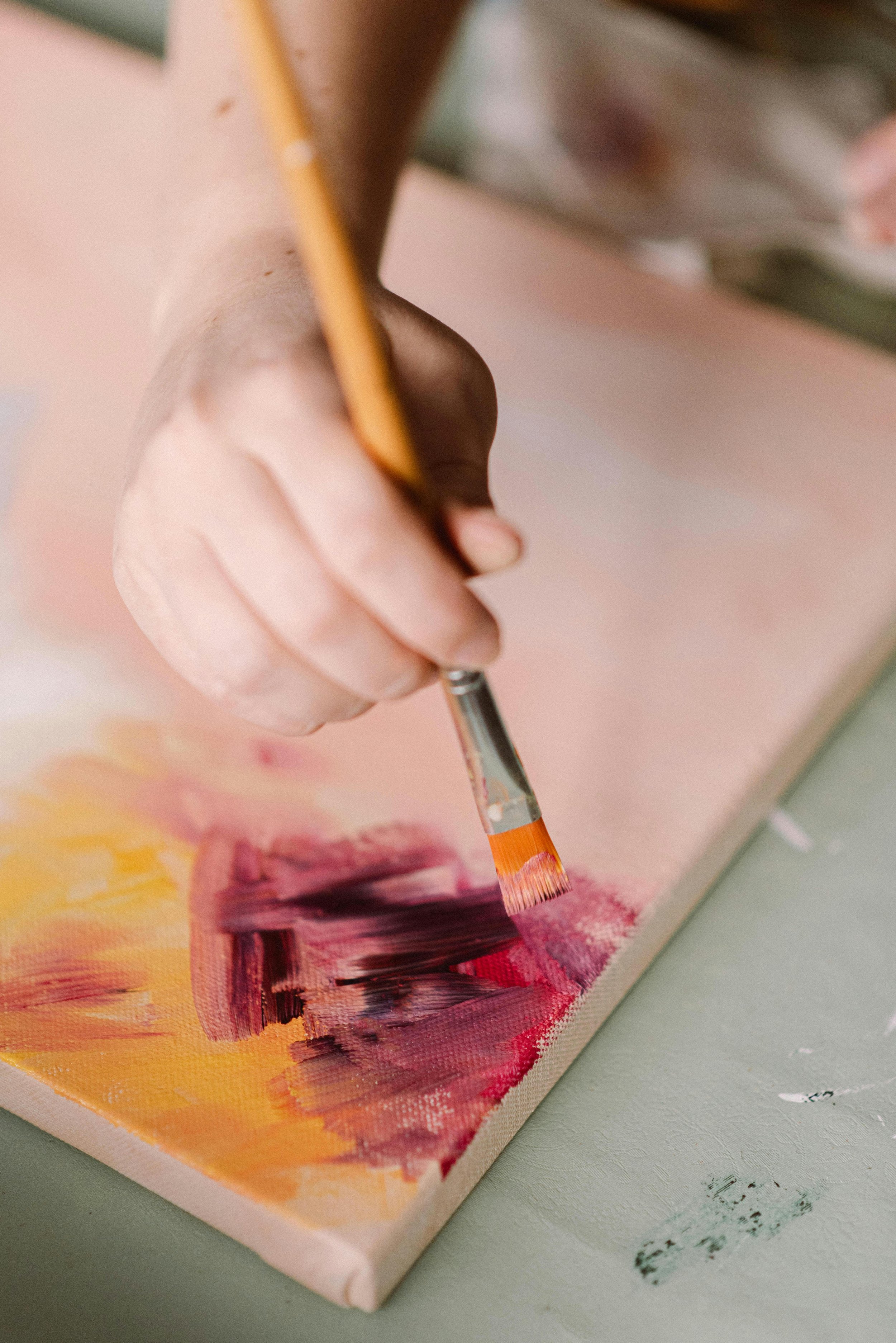 Close-up of a hand holding a paintbrush, applying pink and purple paint on a canvas.