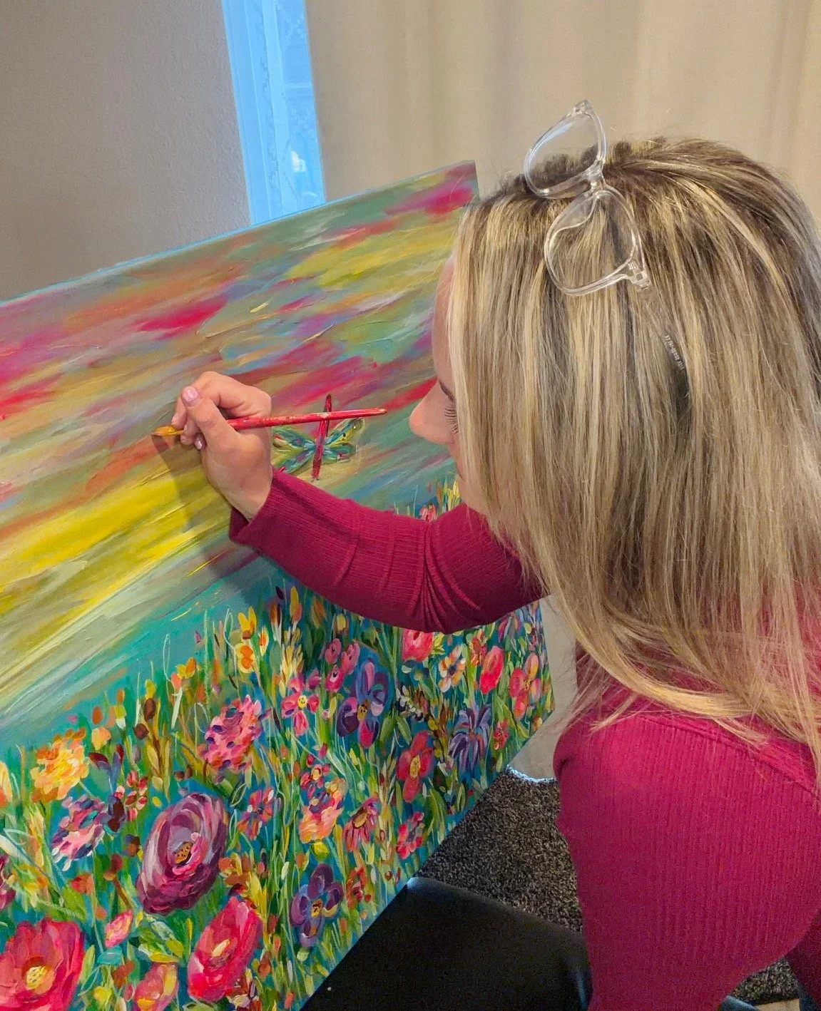 A woman with blonde hair wearing a red long-sleeve shirt, painting a colorful, floral landscape on a large canvas. She has clear glasses resting on her head and is using a red paintbrush.
