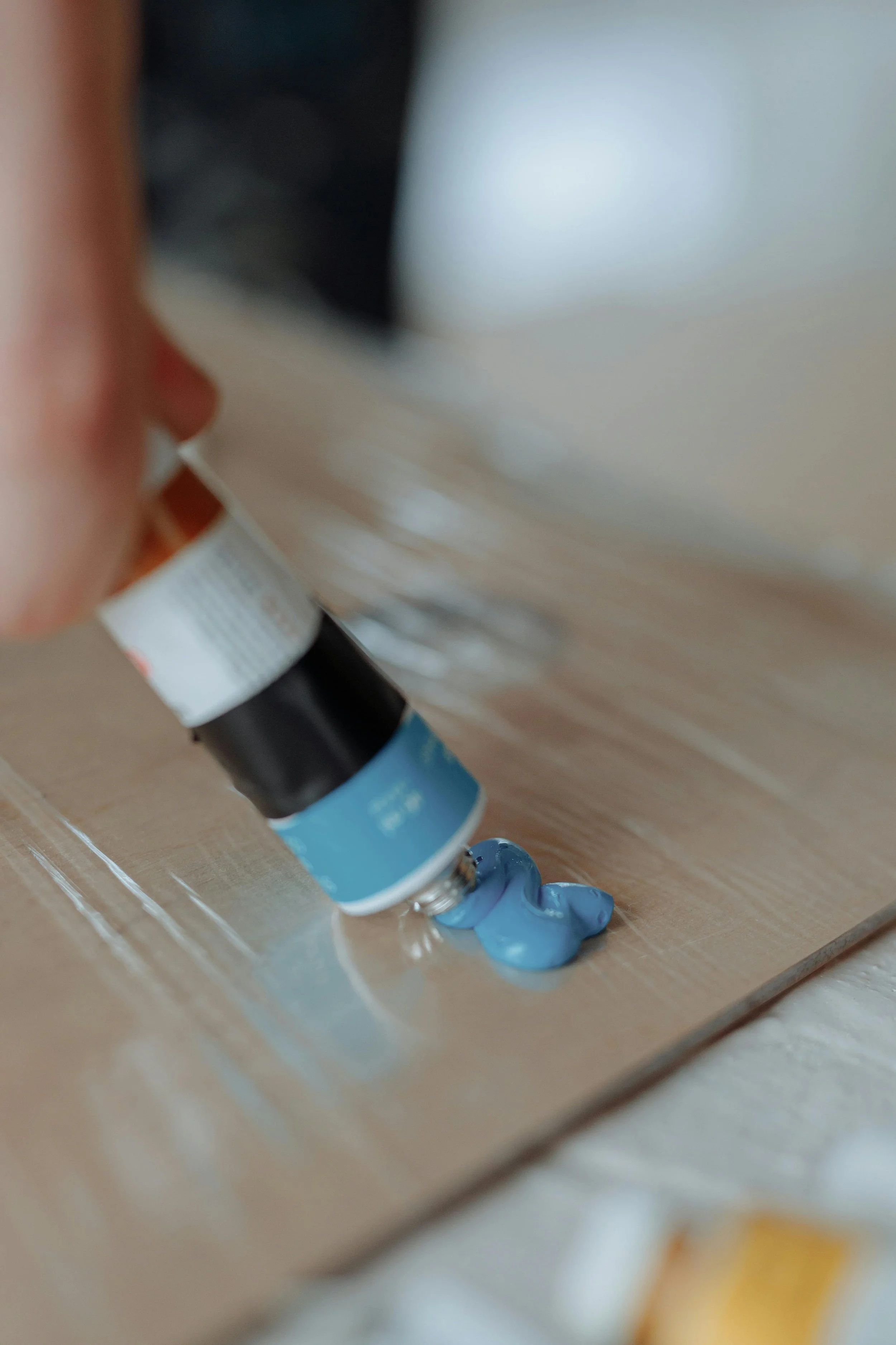 Close-up of a hand squeezing blue paint from a tube onto a wooden surface, with some of the paint spilling out.