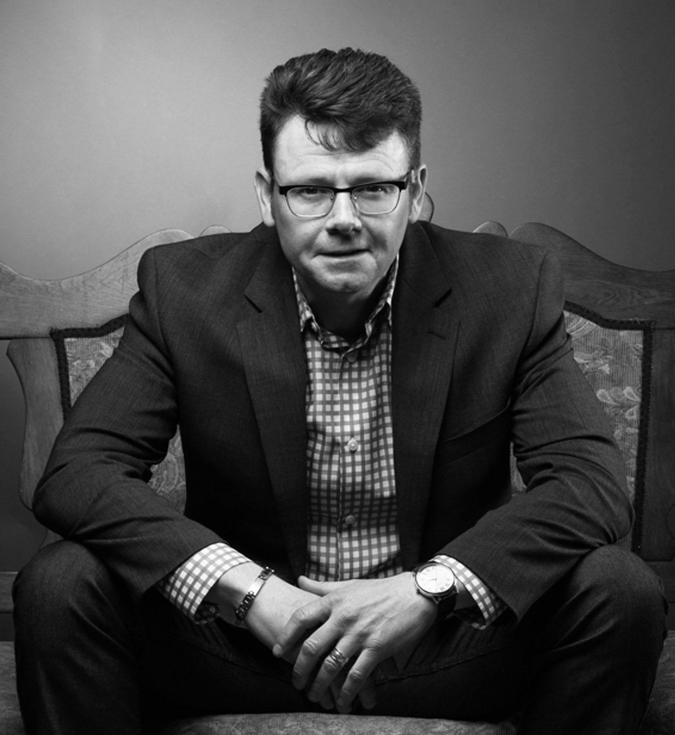 A man in a suit with glasses sitting on a wooden bench with a patterned cushion, wearing a checkered shirt and watch, looking at the camera.
