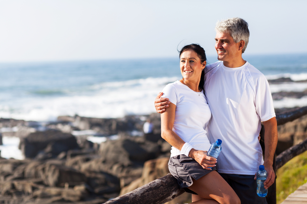 A smiling middle-aged man and woman holding water bottles, standing together by the rocky shoreline at the beach, with ocean waves in the background.