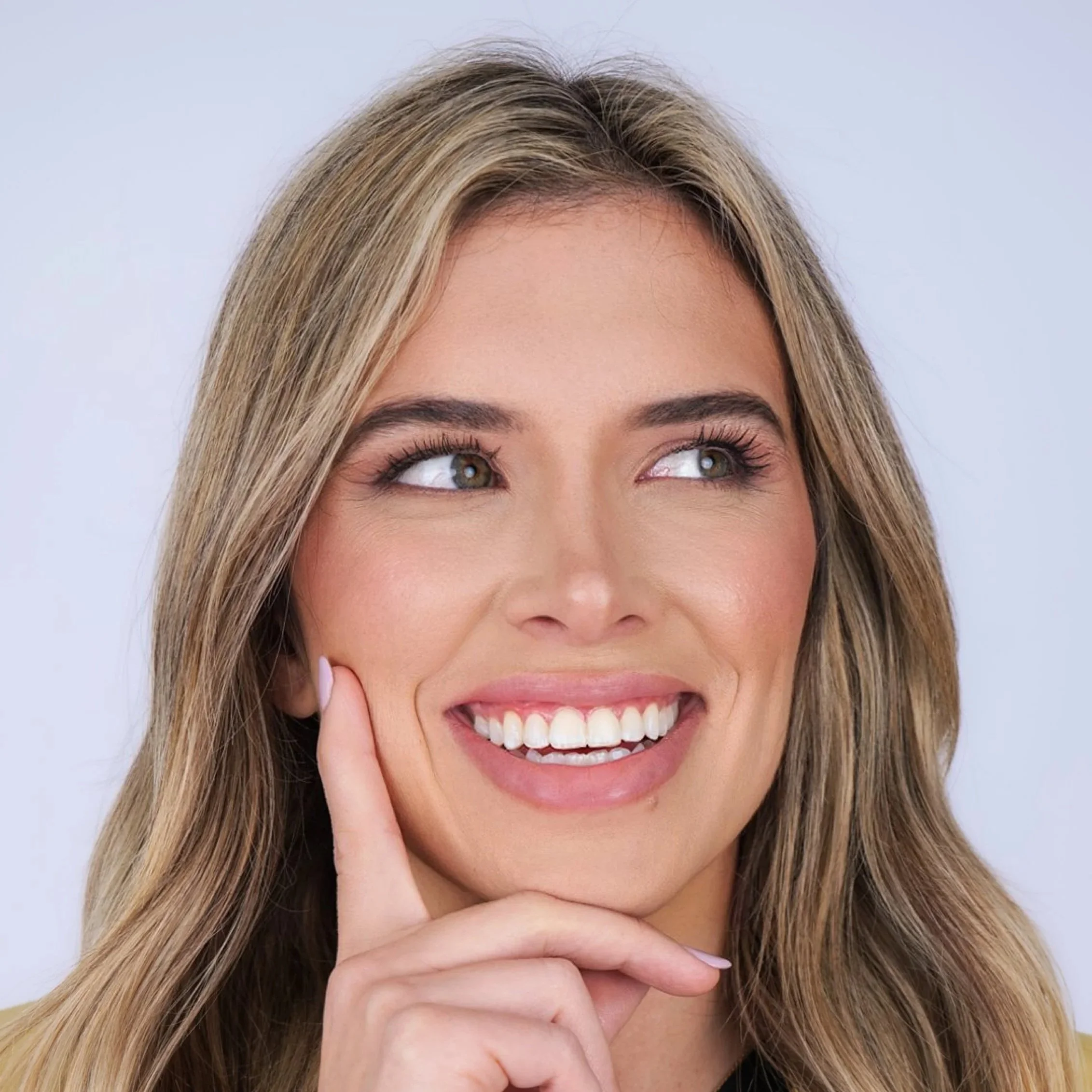 A smiling woman with long blonde hair and an expression of thoughtfulness, touching her face with her index finger.