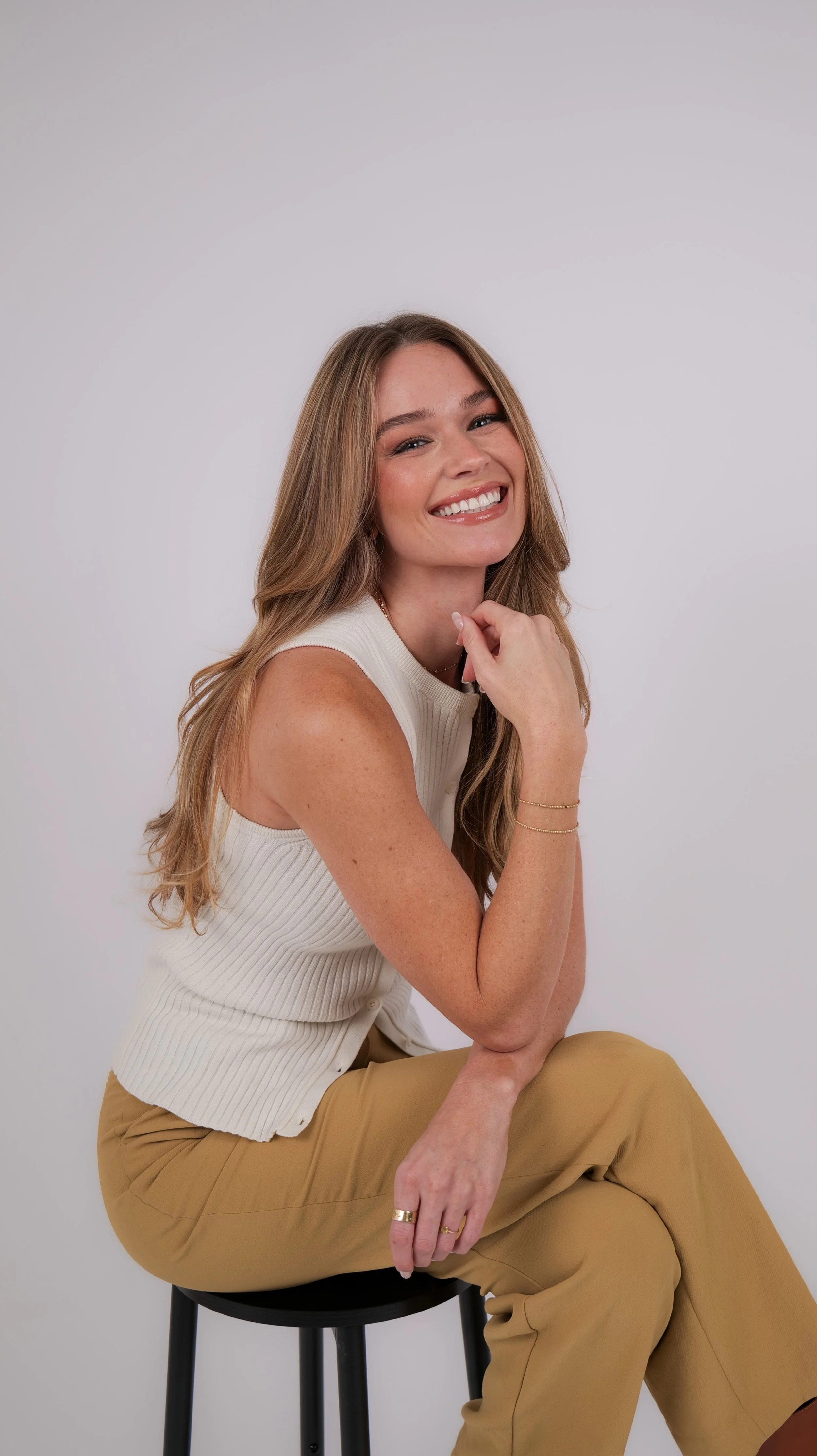 A woman with long blond hair sitting on a black stool, smiling, wearing a sleeveless off-white top and beige pants, against a plain light gray background.