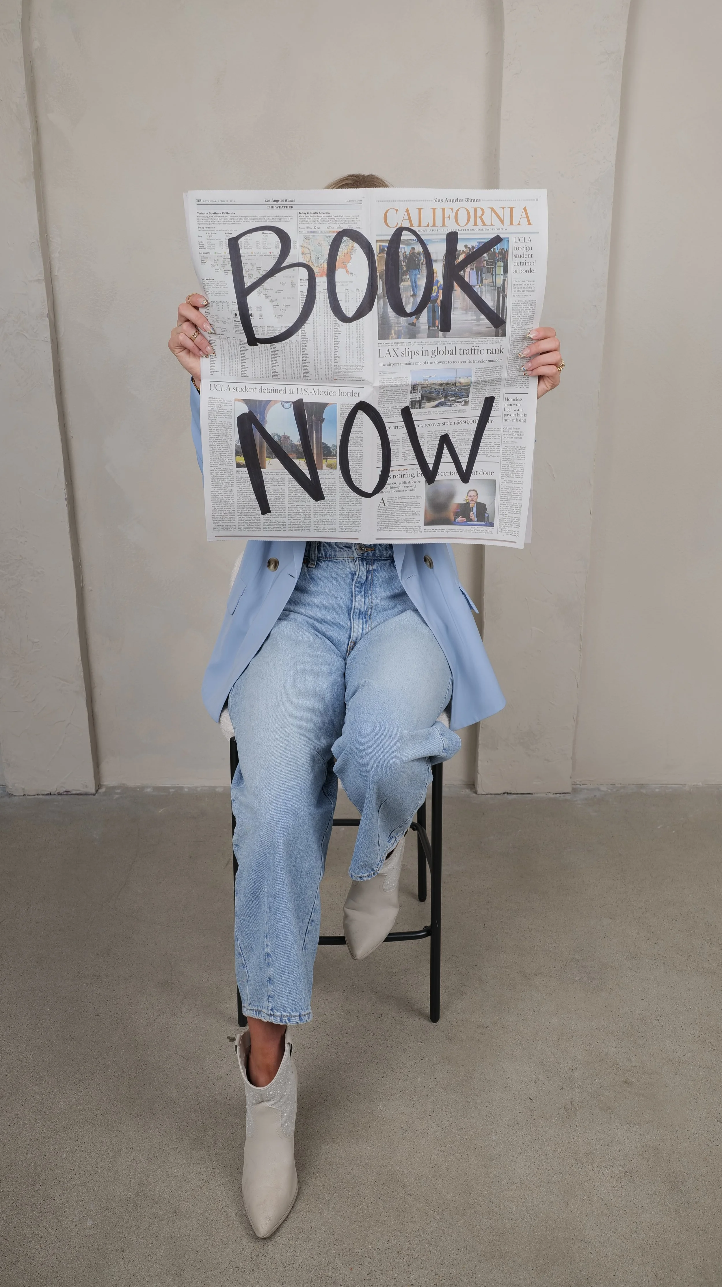 Person sitting on a chair holding a newspaper with 'BOOK NOW' written on it in large black letters, wearing a light blue blazer, jeans, and white boots, against a plain wall background.