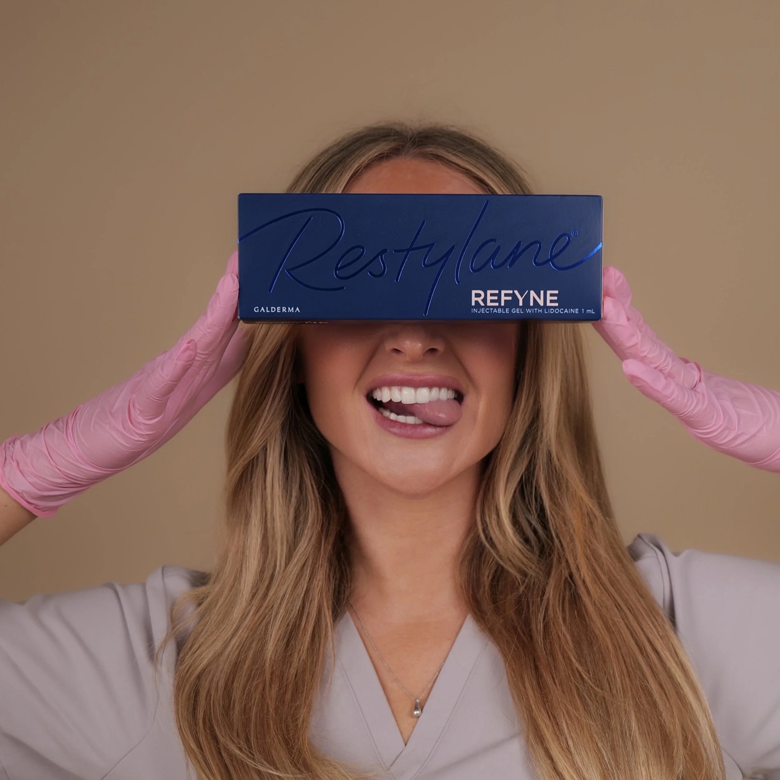 A woman wearing pink medical gloves holds a blue box of Restylane ReFyne in front of her eyes, sticking out her tongue and smiling.