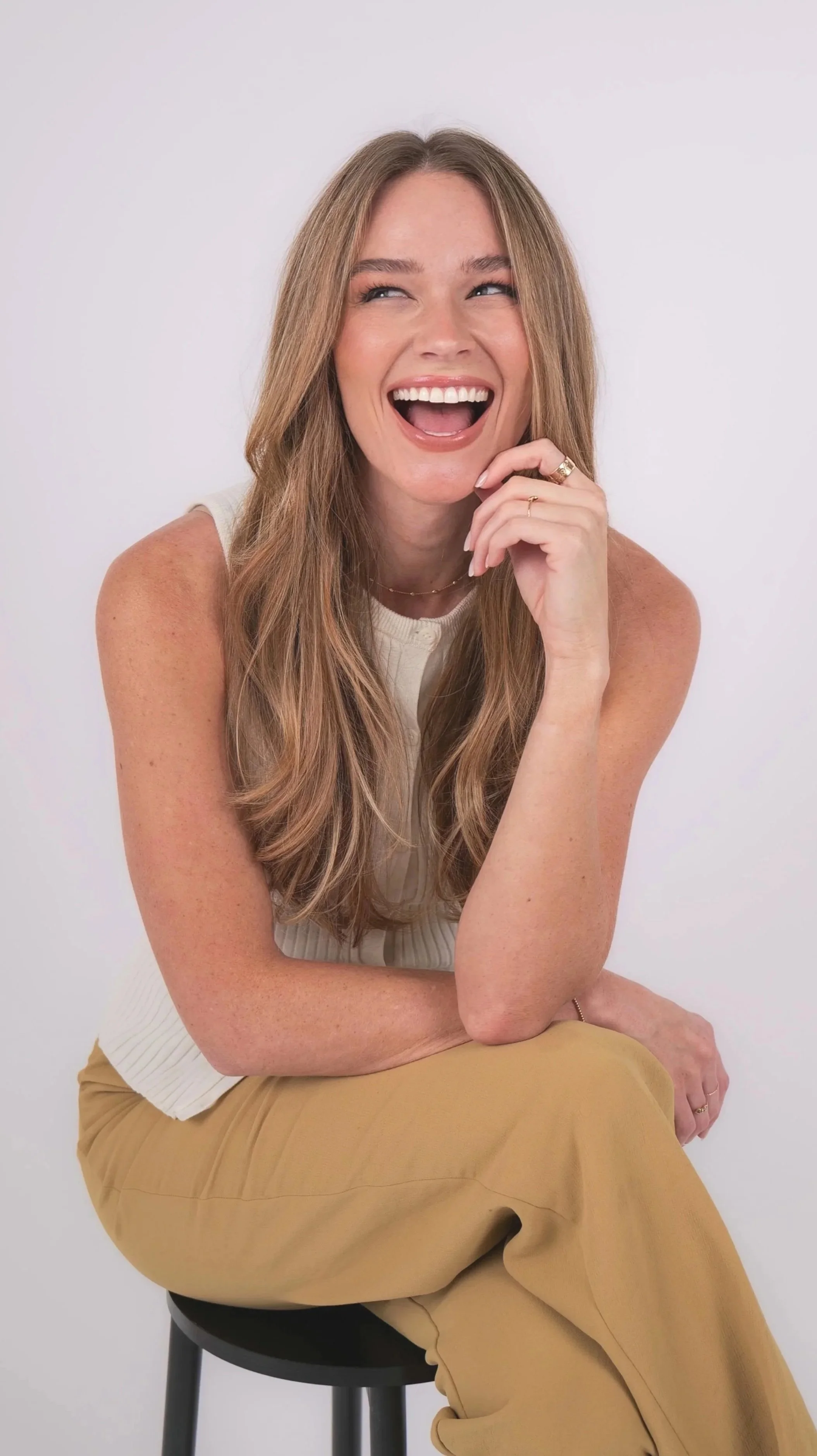 A woman with long red hair, wearing a white sleeveless top and khaki pants, sitting on a black stool against a plain white background, laughing with her mouth open and her hand near her face.