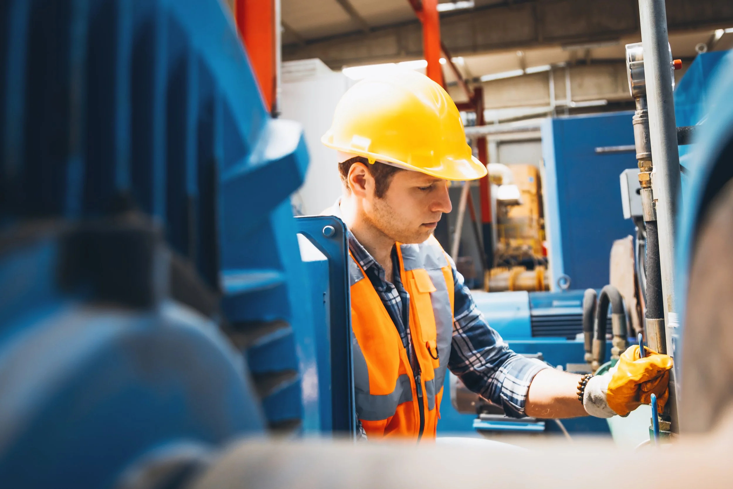 A worker in a yellow safety helmet and orange safety vest working with industrial equipment in a factory or workshop.