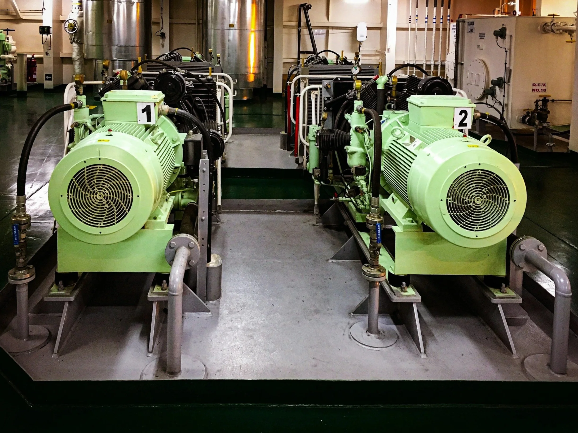 Two large green industrial compressors with black hoses connected, labeled with numbers 1 and 2, in a factory or mechanical room with metallic tanks and other equipment in the background.