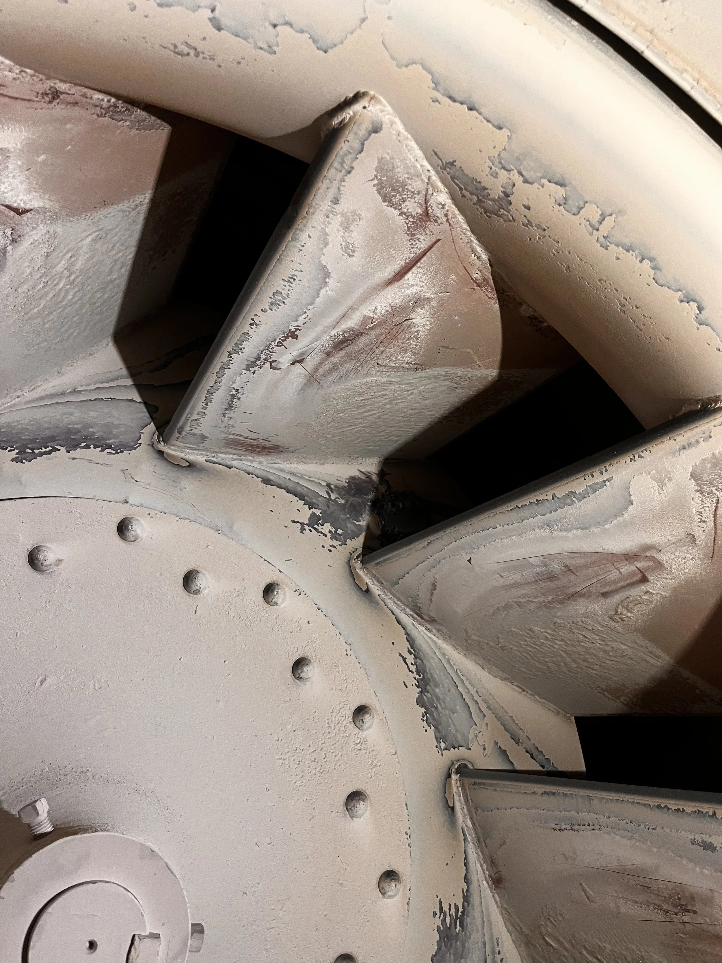 Close-up of a worn, weathered metallic surface with peeling paint, dents, and scratches. Parts of the surface are rounded and some have rivets, indicating an industrial or mechanical object.