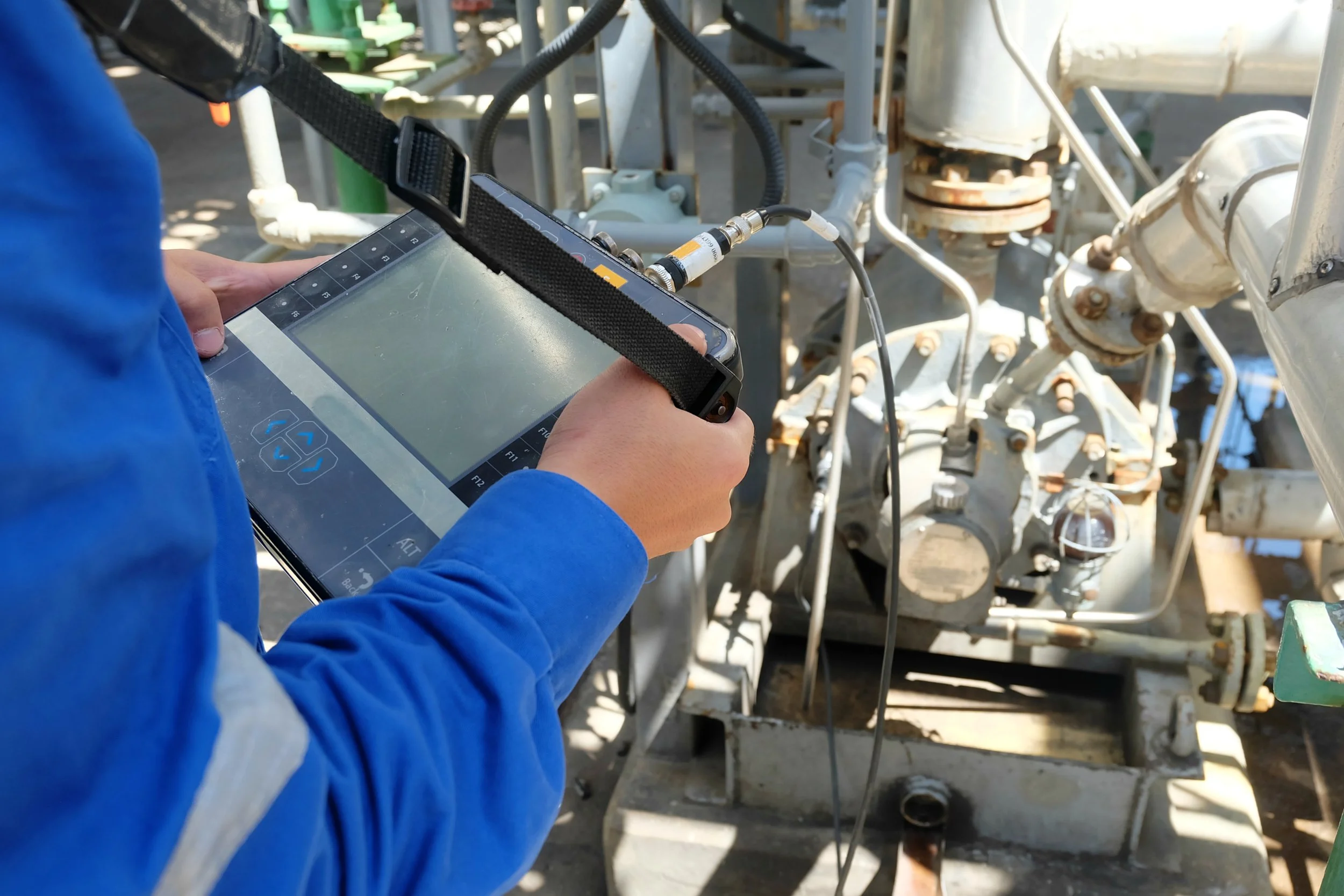 A worker in a blue jacket holding a handheld device connected to industrial equipment with black straps, performing tests or diagnostics on machinery outside.