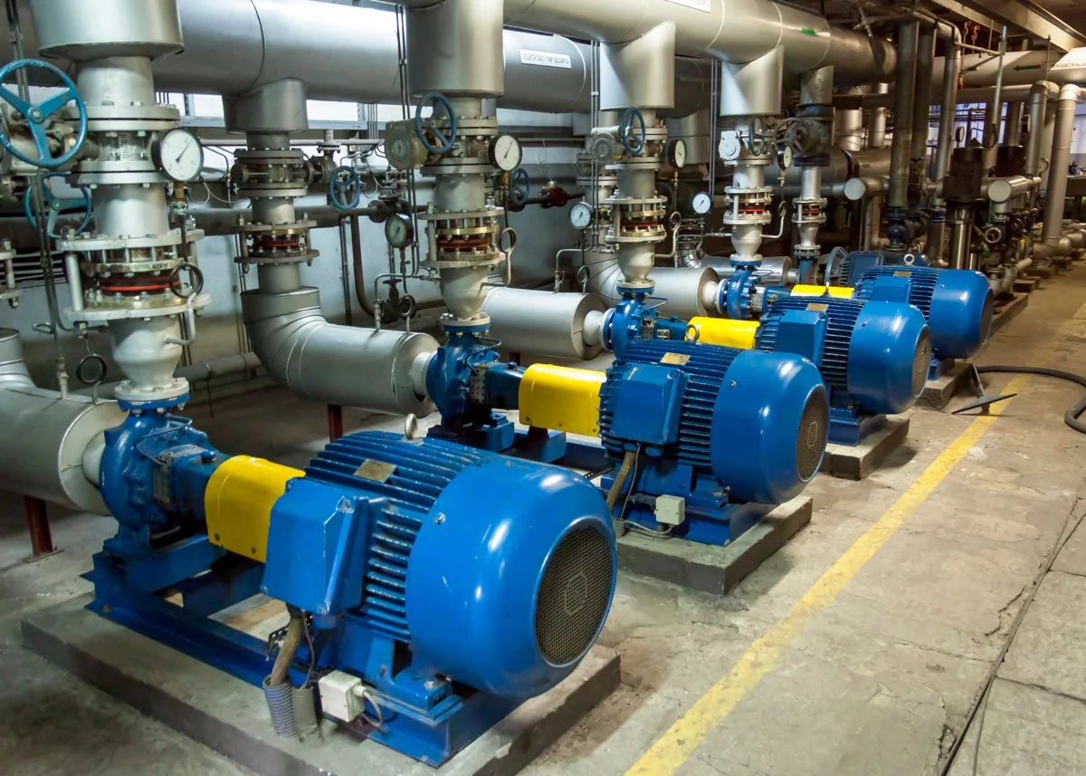 Industrial pump room with four large blue pumps connected to metallic piping, gauges, and valves.