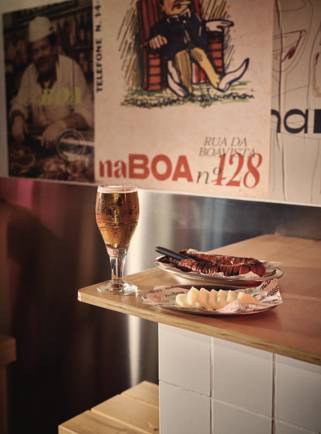 Taça de vinho rosé, prato com queijo e calabresa, e uma parede com pôster antigo.