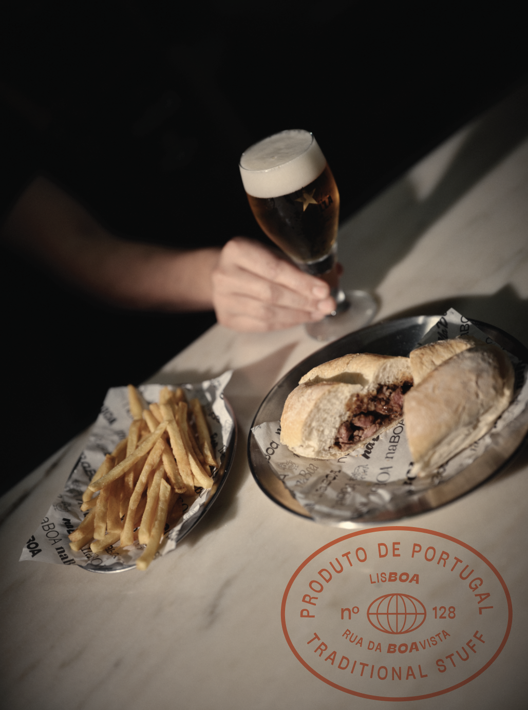 Pessoa segurando taça de cerveja enquanto come um sanduíche de carne e batatas fritas em um restaurante.