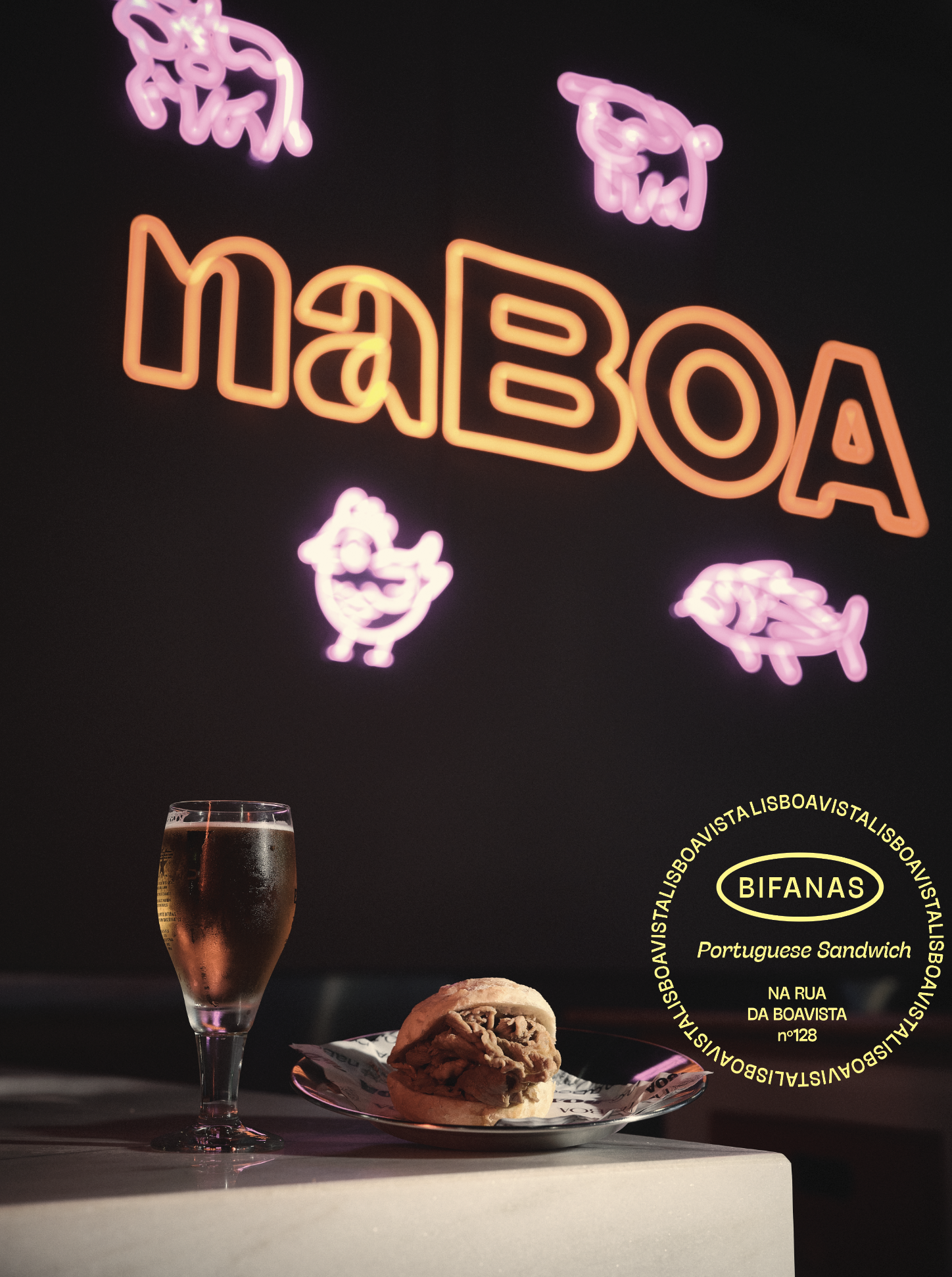 Decoração com letreiro de neon com a palavra 'Maía' na cor laranja e desenhos de pintainhos e patinhos em neon rosa, com uma taça de espumante, um pão recheado e uma embalagem de pão na mesa.