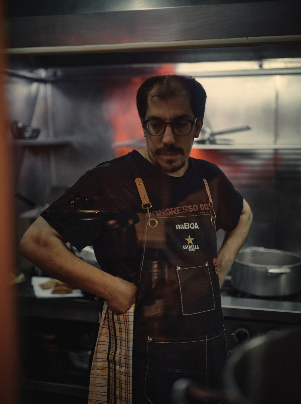 Homem com óculos e barba, usando avental, em uma cozinha industrial, olhando para baixo com as mãos na cintura.